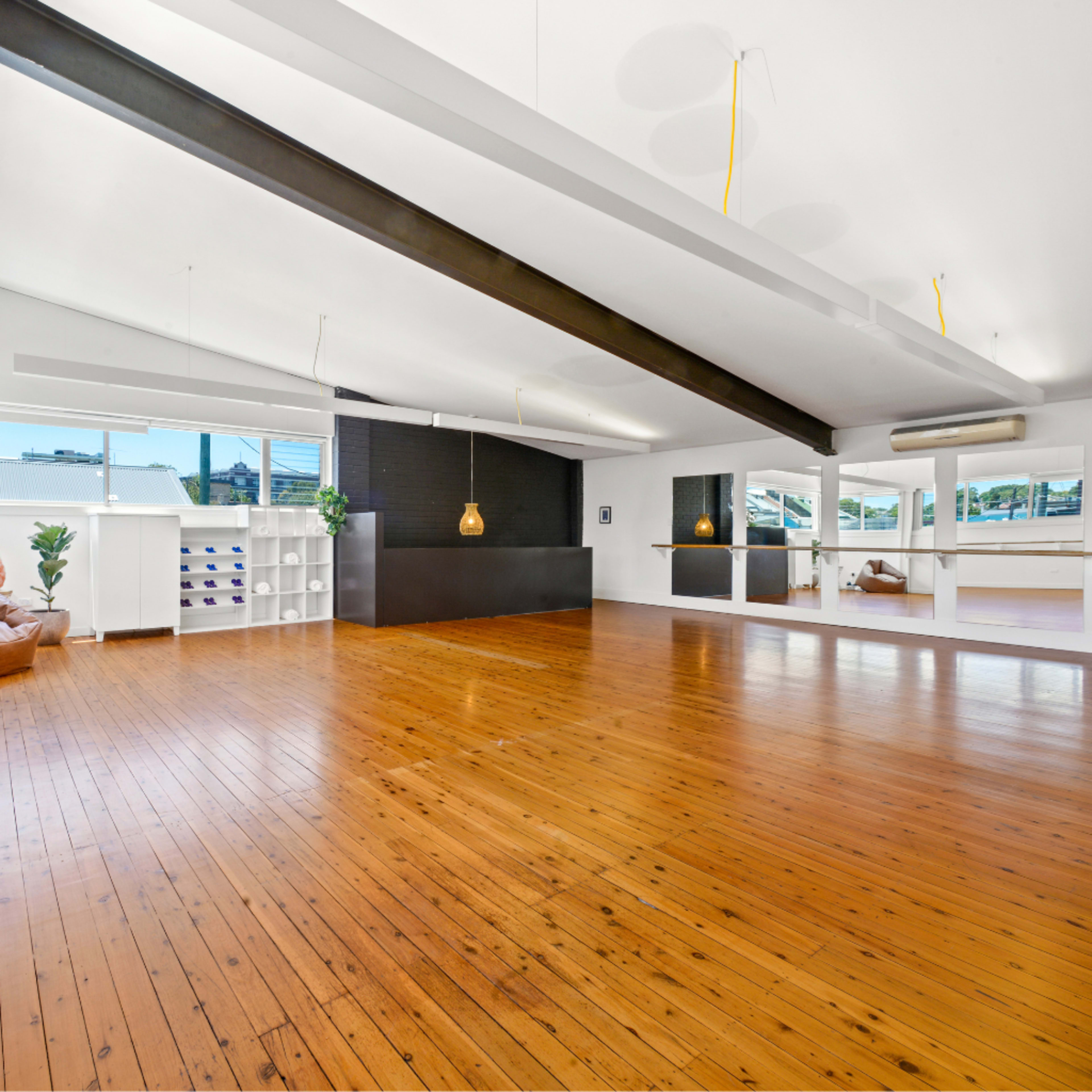A spacious, light-filled room with wooden flooring, large windows, and a minimalistic design including a seating area and a black feature wall.