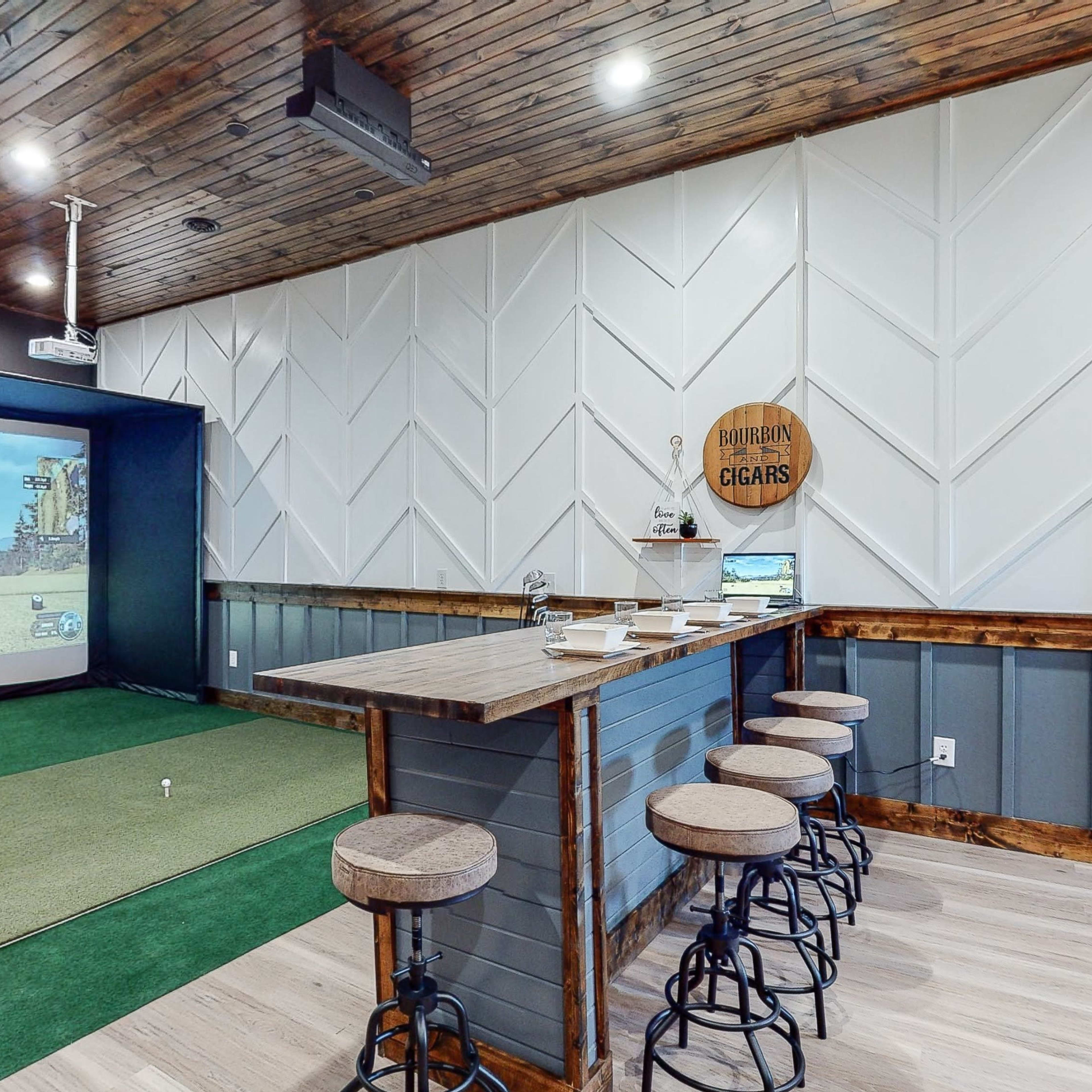 An indoor golf simulation room featuring a large projected screen, a green turf area, and a bar with stools.