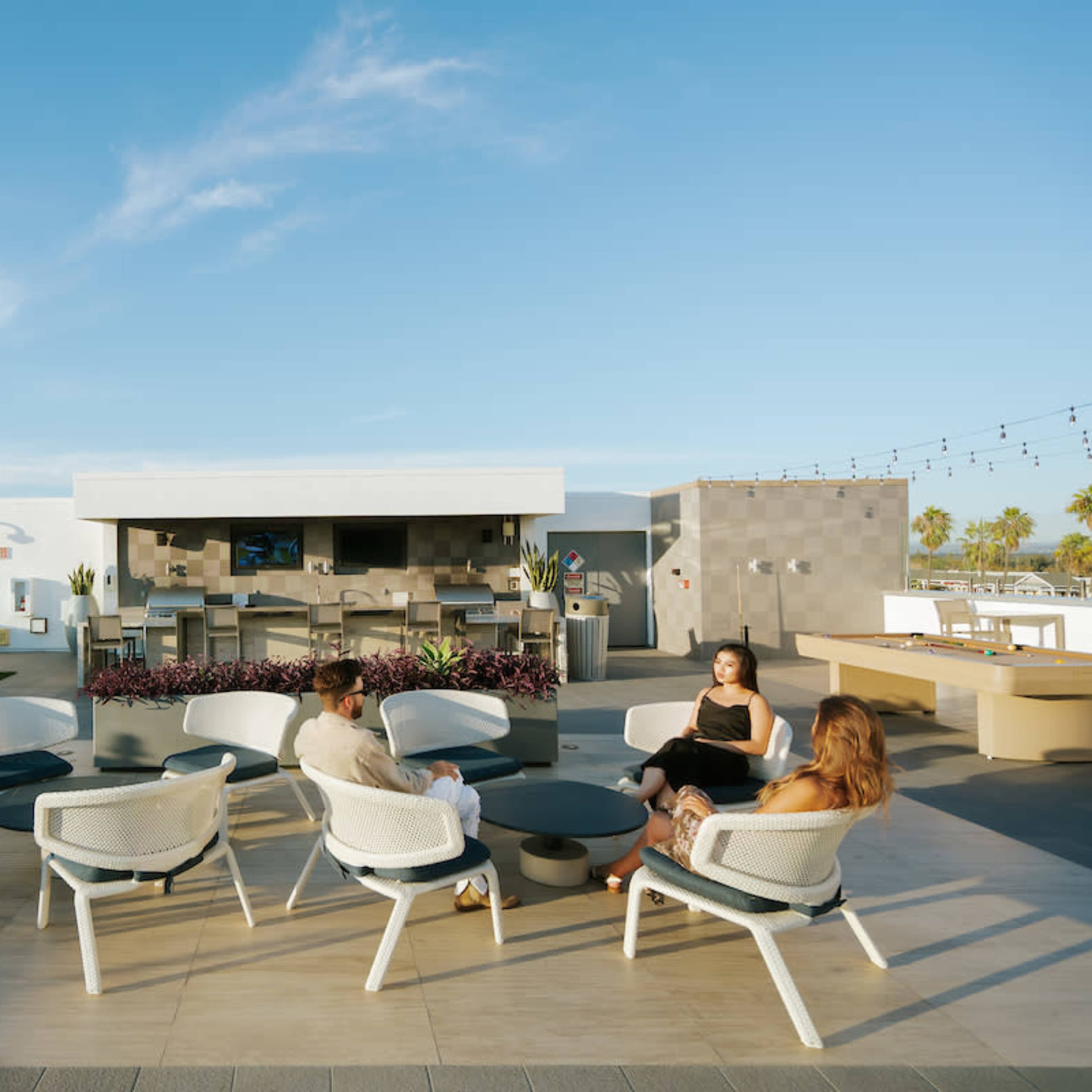 A group of people sits around a circular table on a rooftop patio with a bar and lounge chairs in the background.
