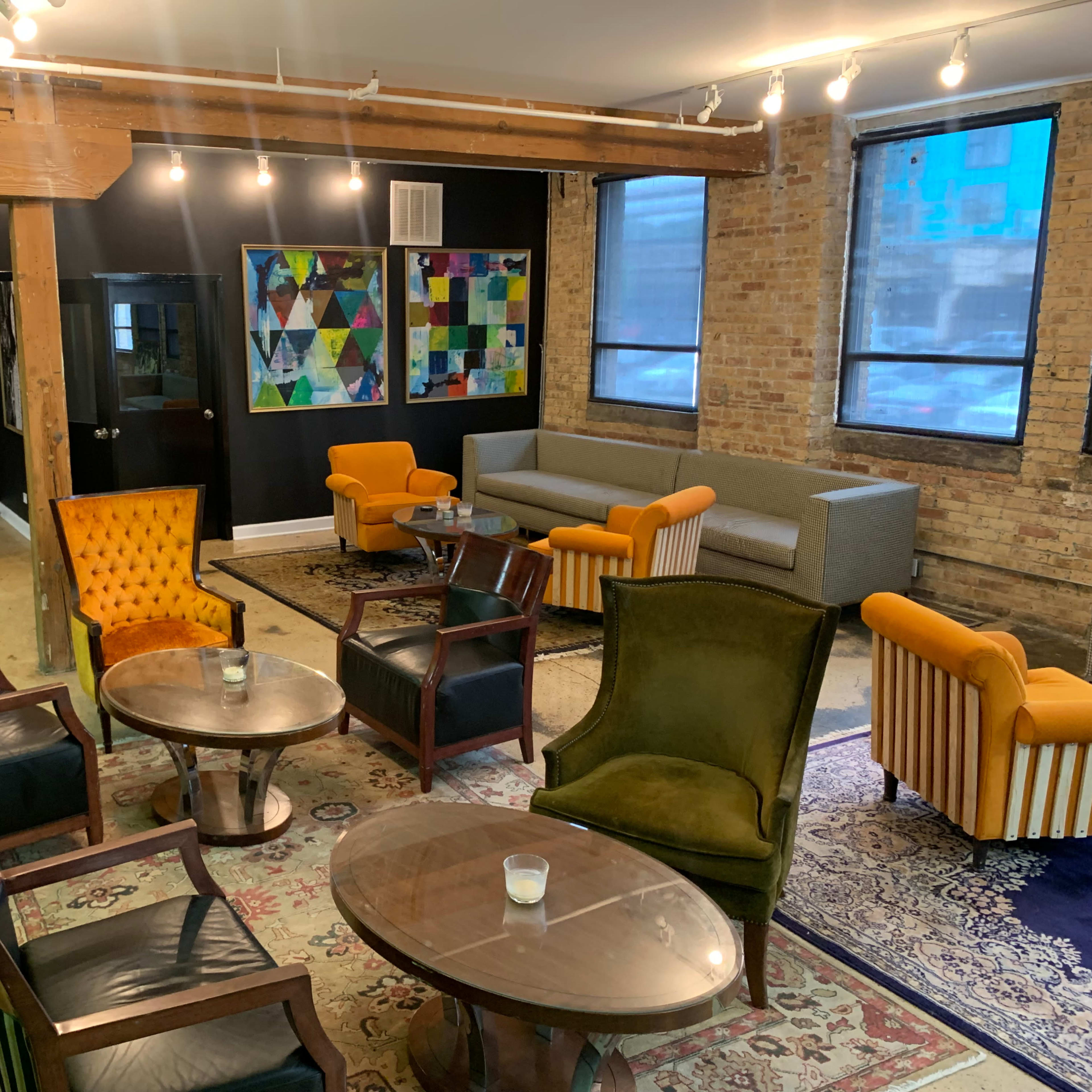 A modern lounge area with a mix of seating arrangements, including orange chairs, a gray sofa, and various tables on a patterned rug.