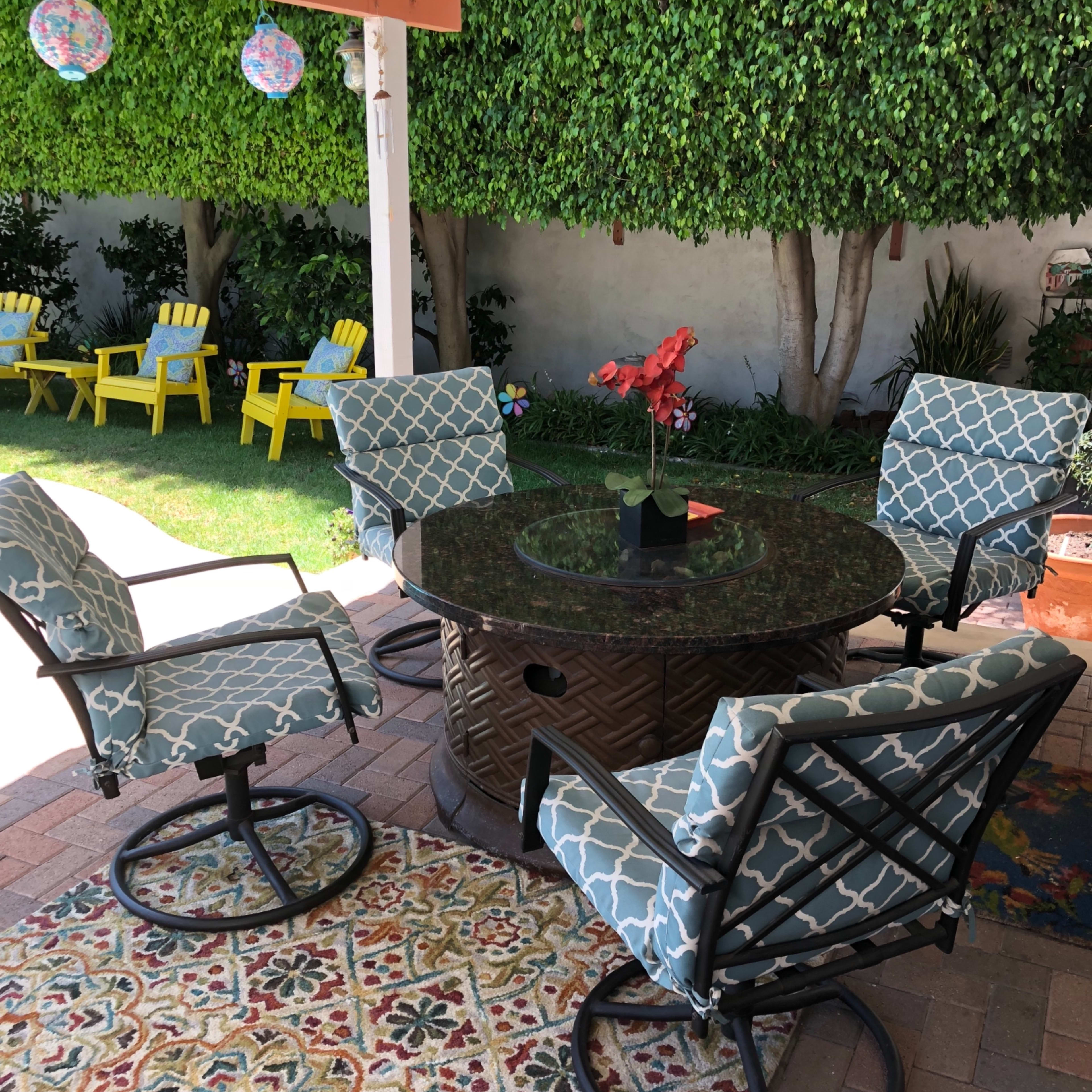 A round table with four patterned chairs sits under a covered patio surrounded by green hedges and a colorful outdoor rug.