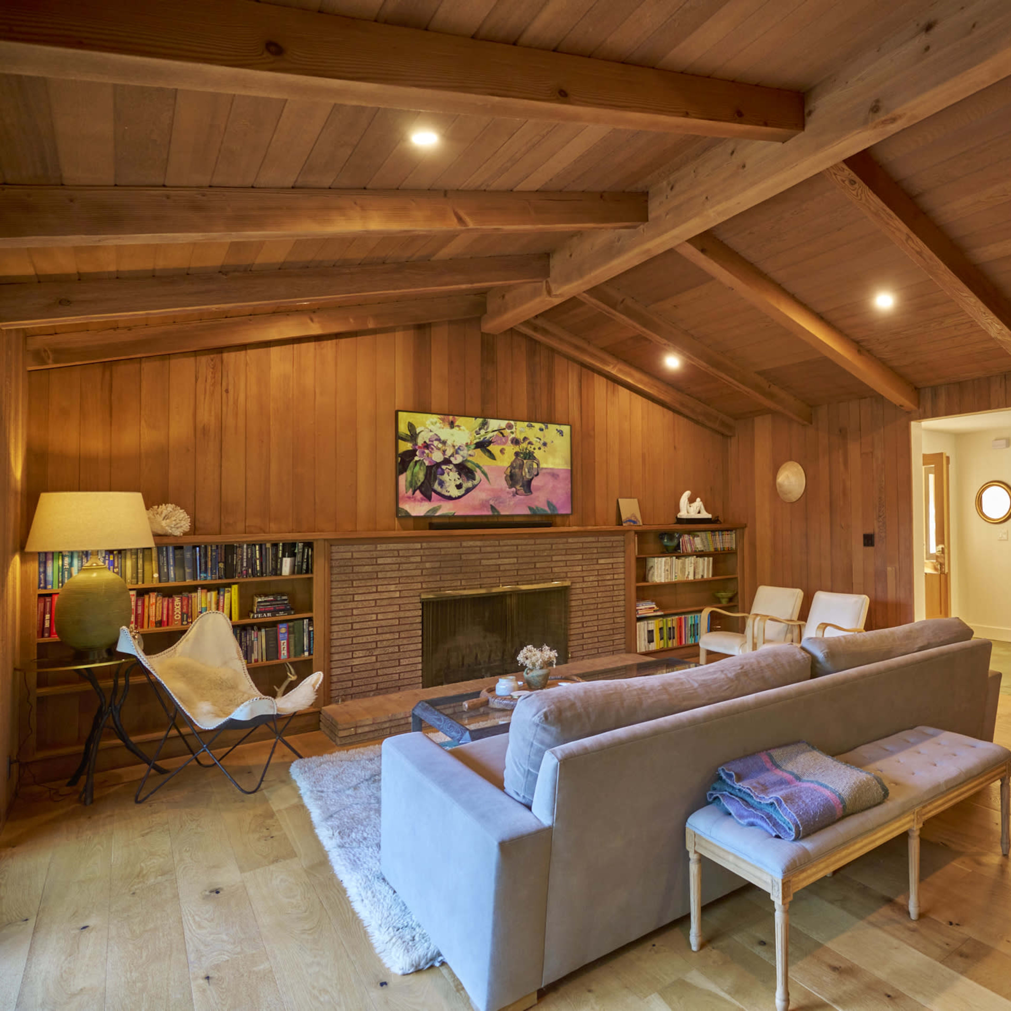 The living room features wooden walls and a ceiling with exposed beams, a brick fireplace, a light gray sofa, and a bookshelf, creating a cozy space.