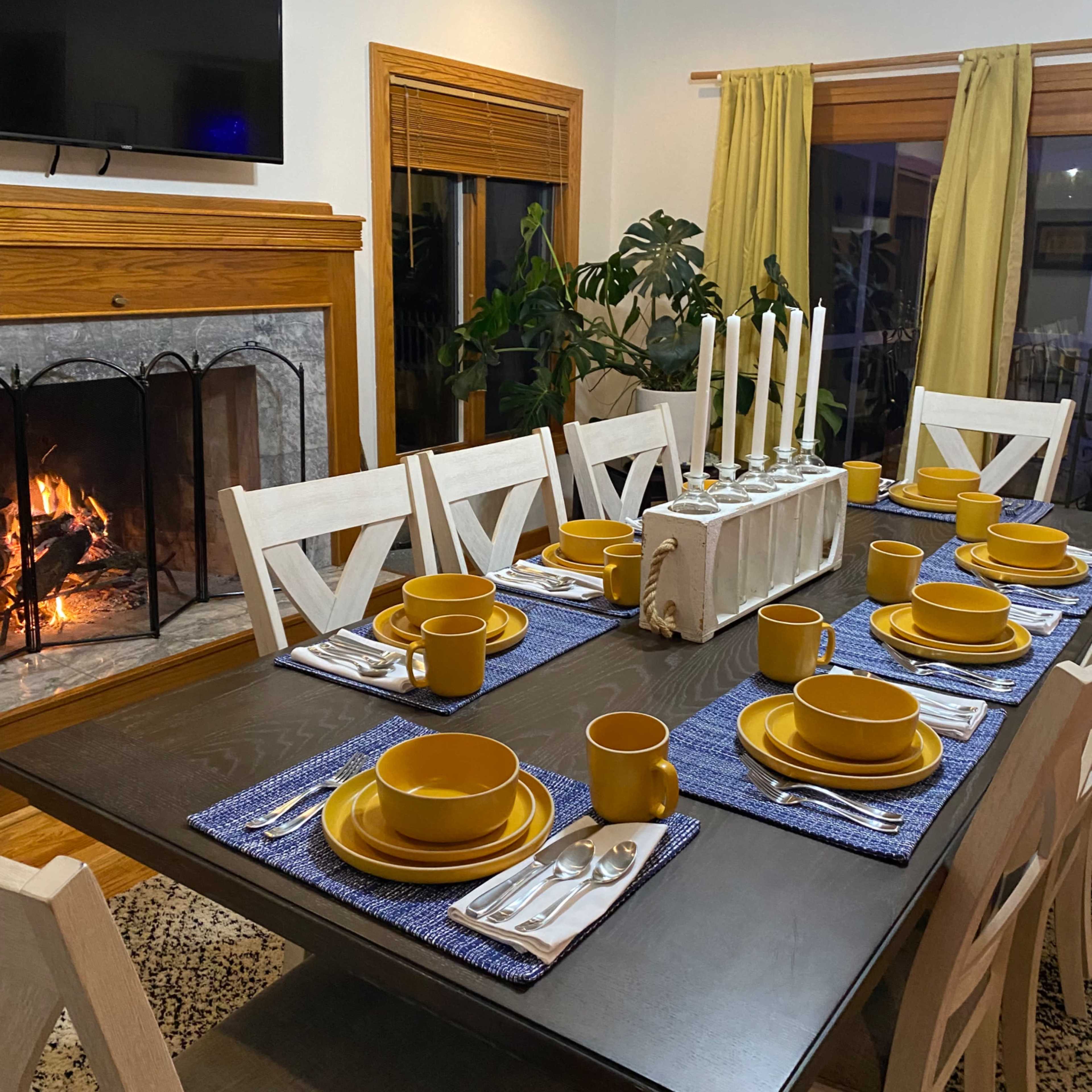 A dining table is set with yellow plates and cups on blue placemats, while a fireplace burns in the background.