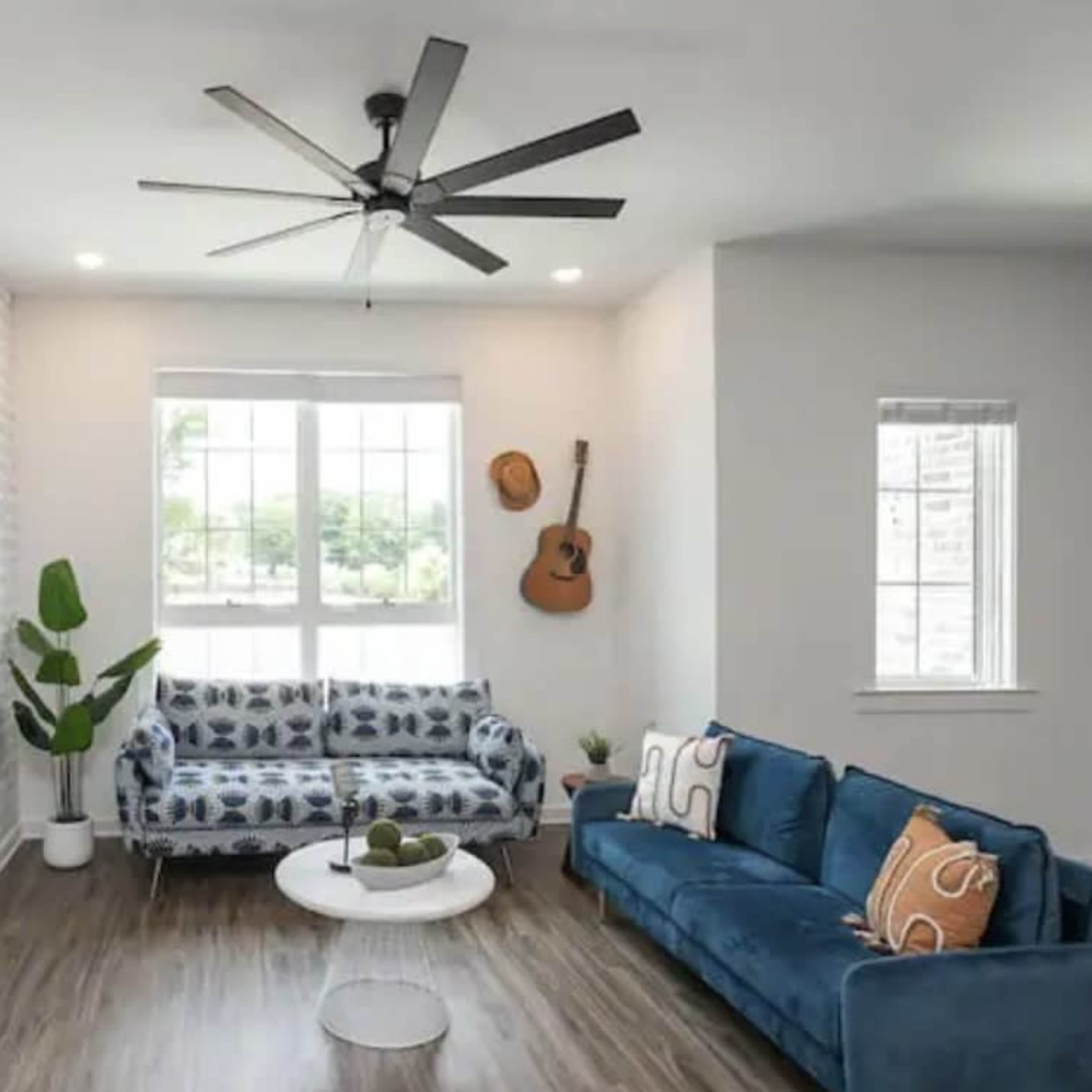 A modern living room features a blue couch, a patterned sofa, a flat-screen TV, and a guitar hanging on the wall, with a large window allowing natural light.