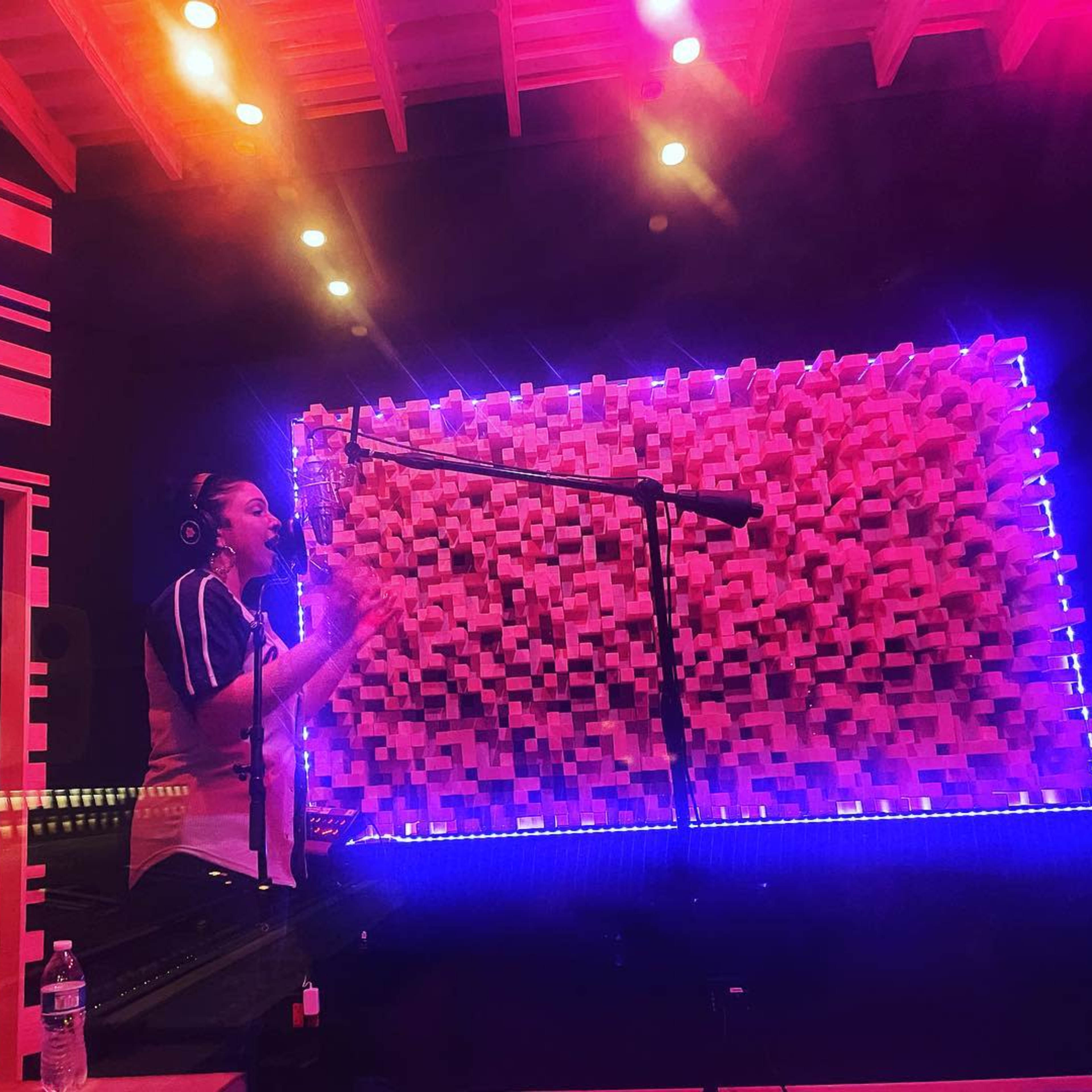 A person is recording vocals in a sound studio with colorful lighting and acoustic wall paneling.