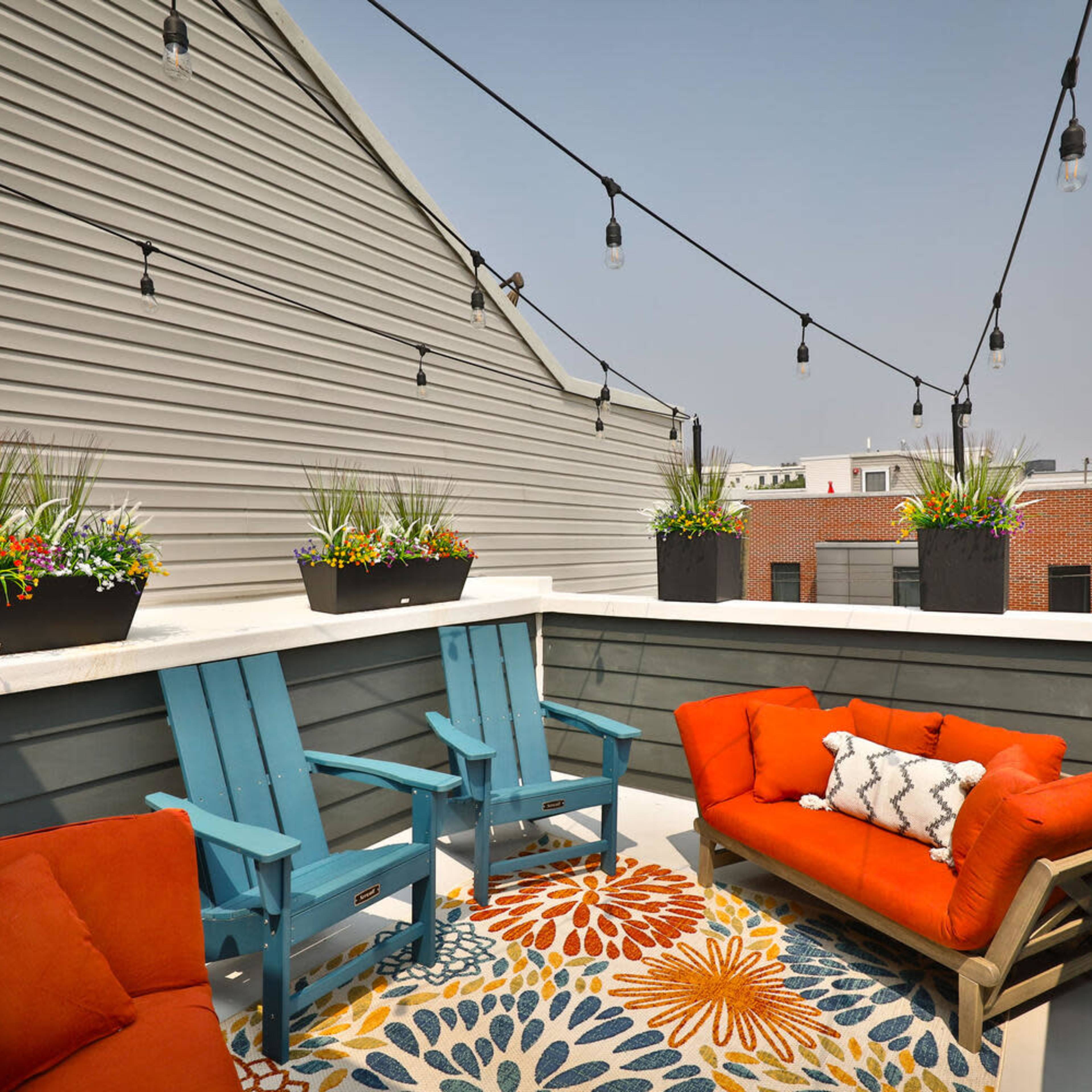 A rooftop patio features orange furniture, a colorful rug, and potted plants, with string lights above.