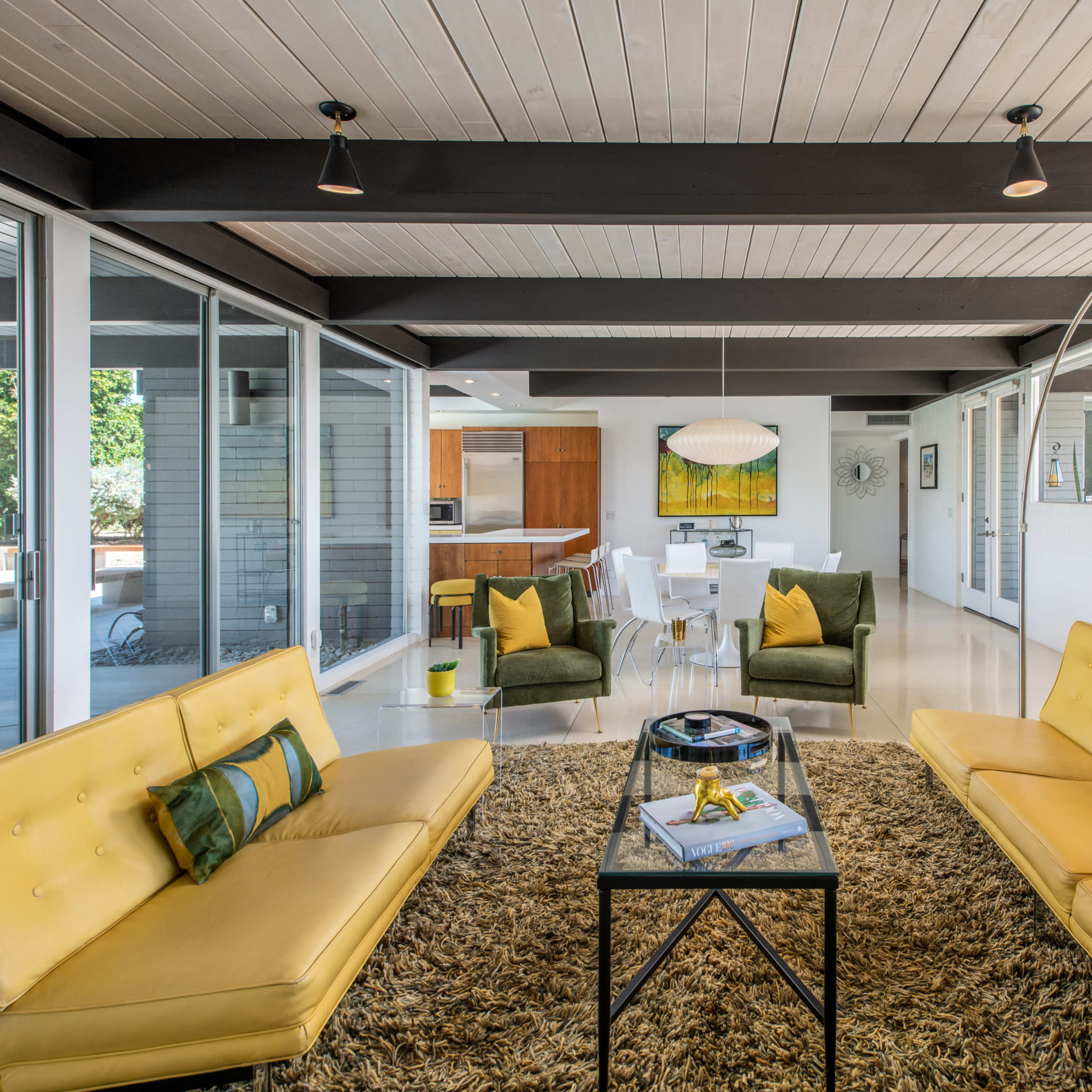 A modern living room with two yellow sofas facing a glass coffee table, surrounded by green armchairs and large windows letting in natural light.