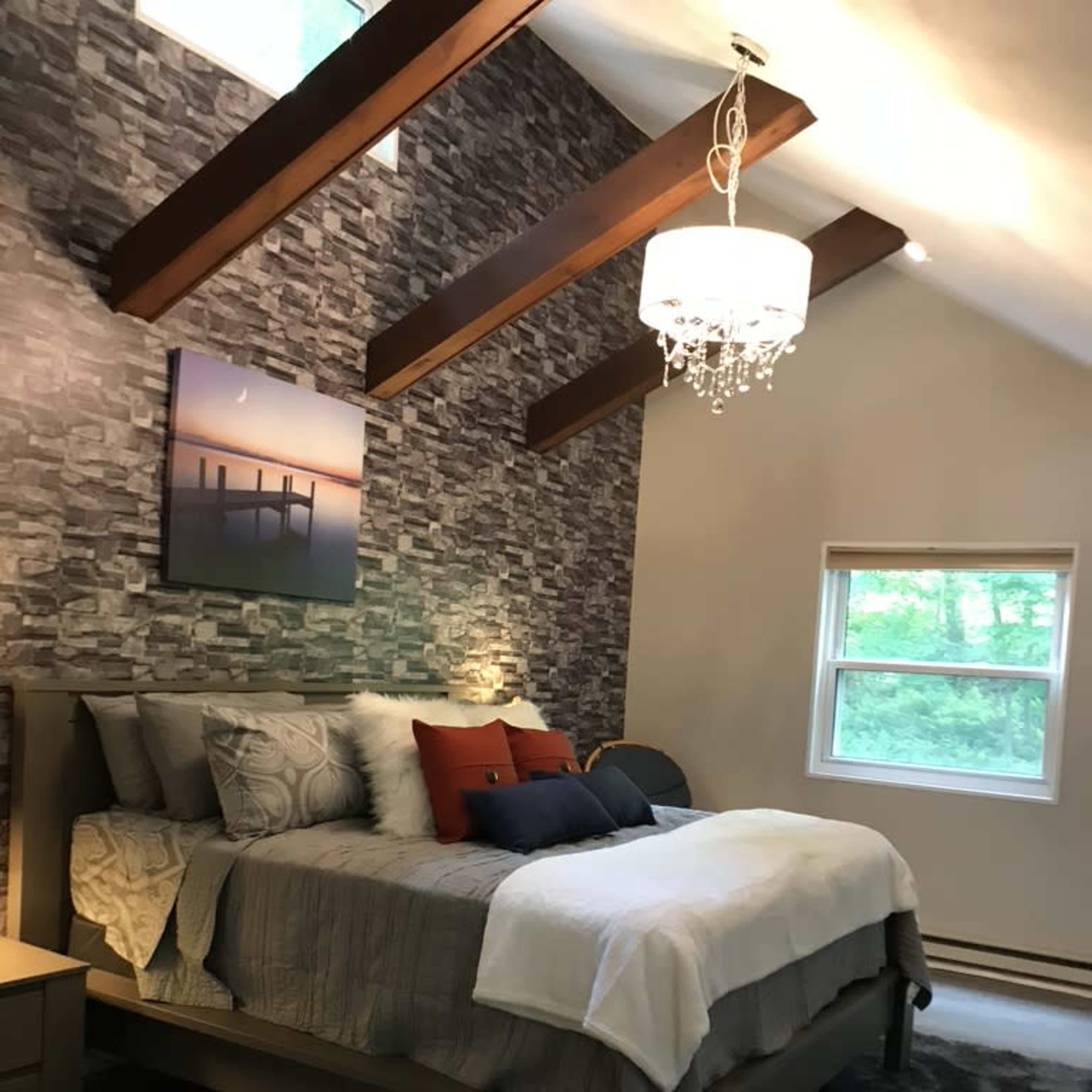 A bedroom with a large bed, a chandelier, and stone accent walls, illuminated by natural light from a nearby window.