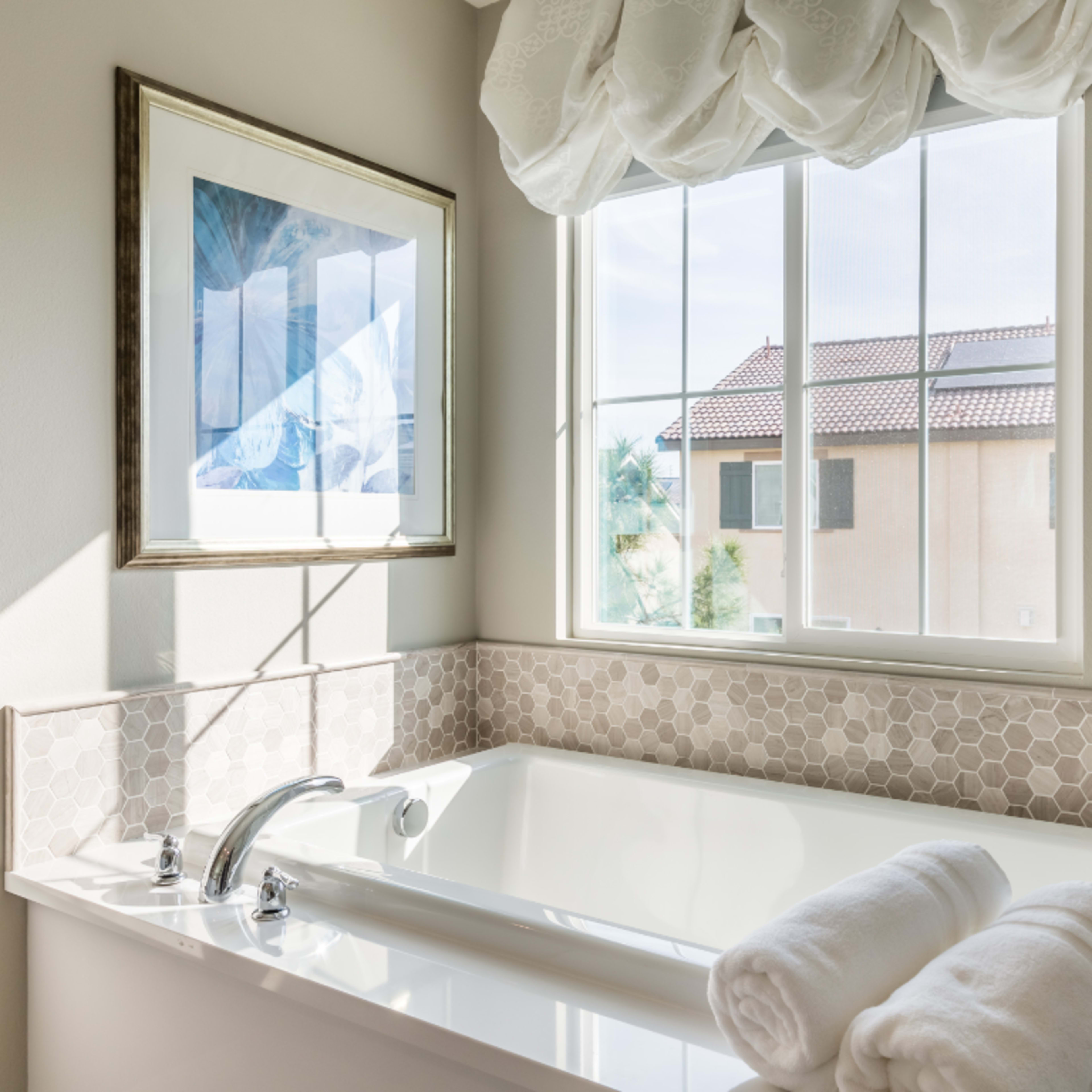 A bright bathroom features a white tub with two rolled towels and a window that provides a view of nearby buildings.