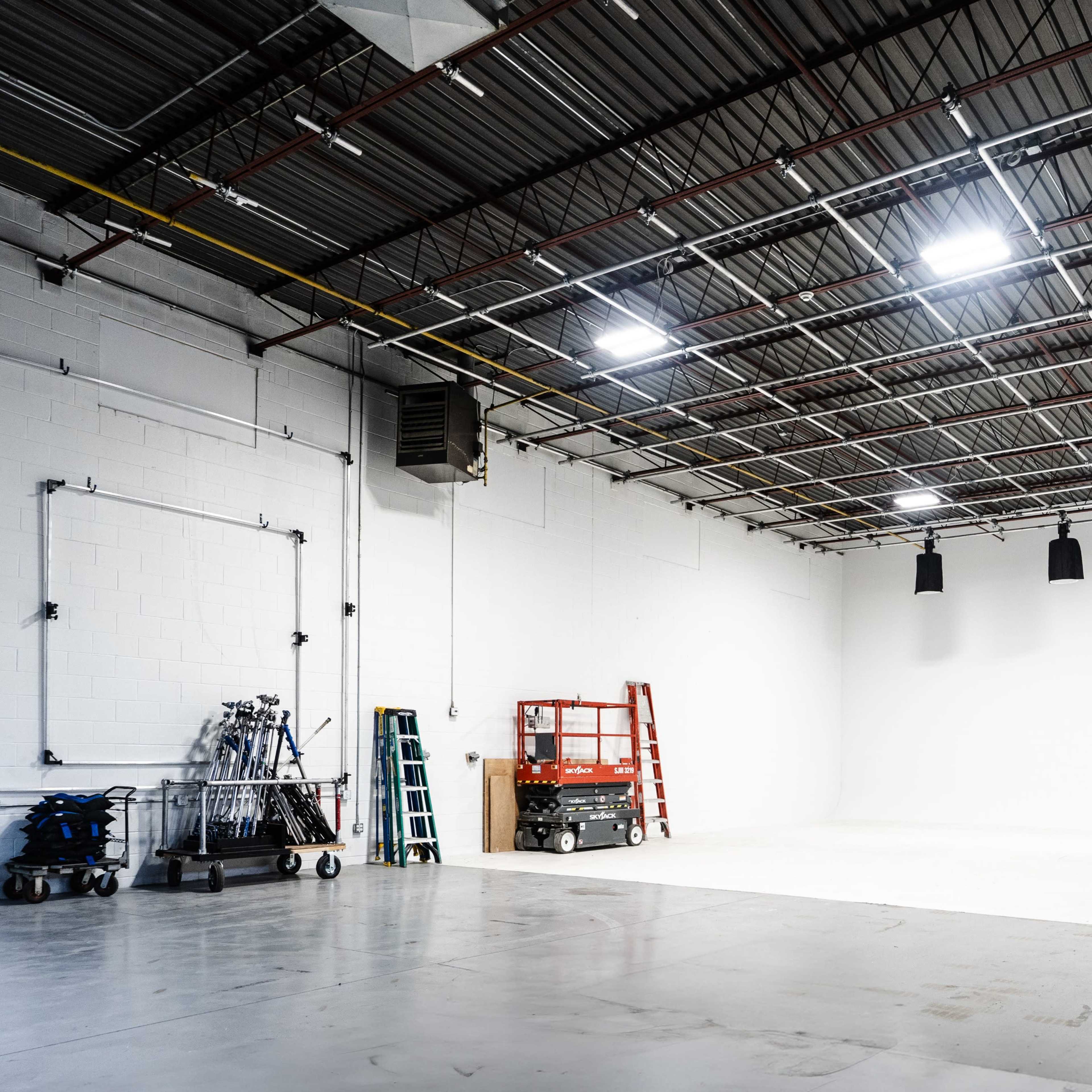 An empty photography studio features a large white backdrop, several ladders, and various lighting equipment along the walls.