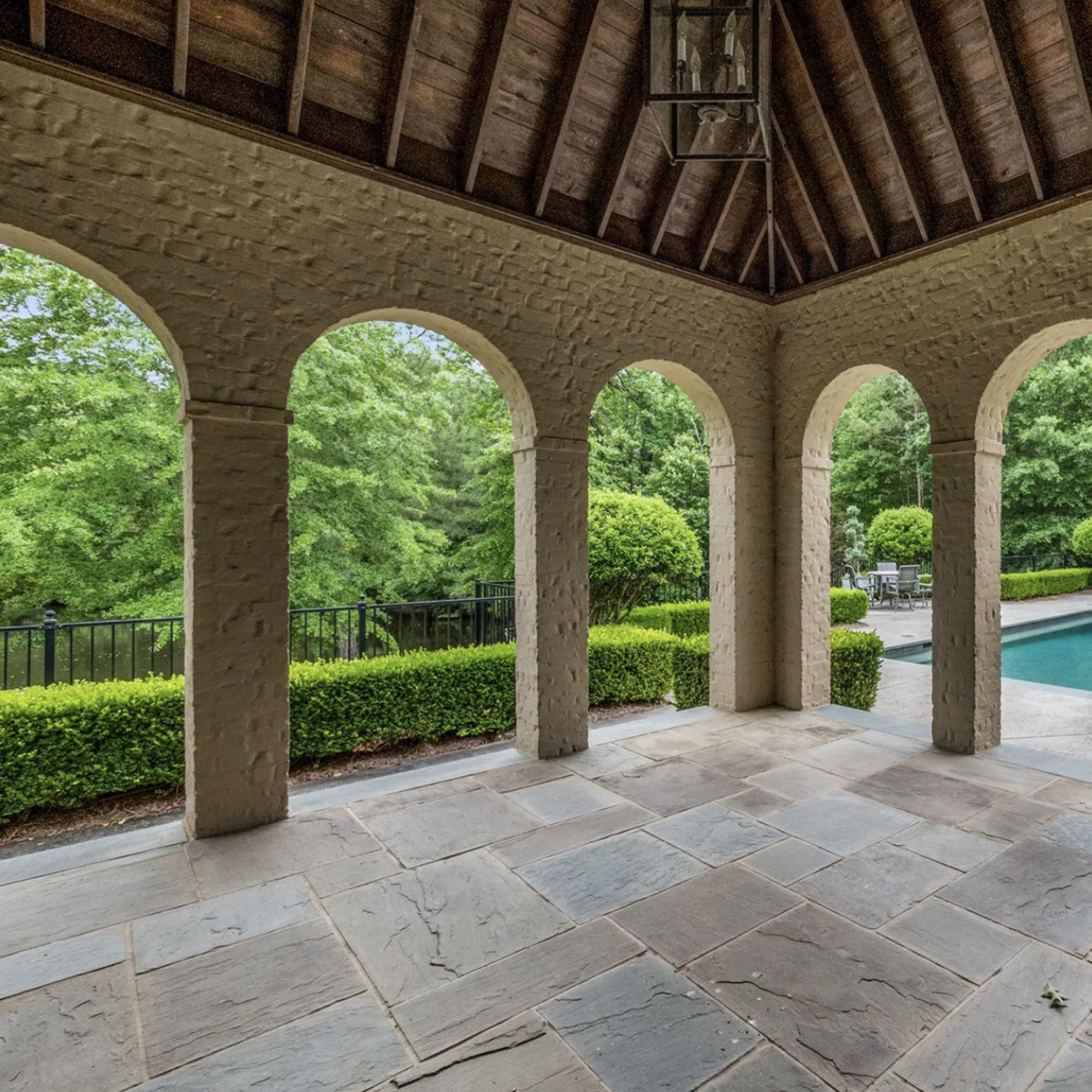 A covered outdoor patio with arched openings, stone flooring, and a view of a landscaped pool area surrounded by greenery.