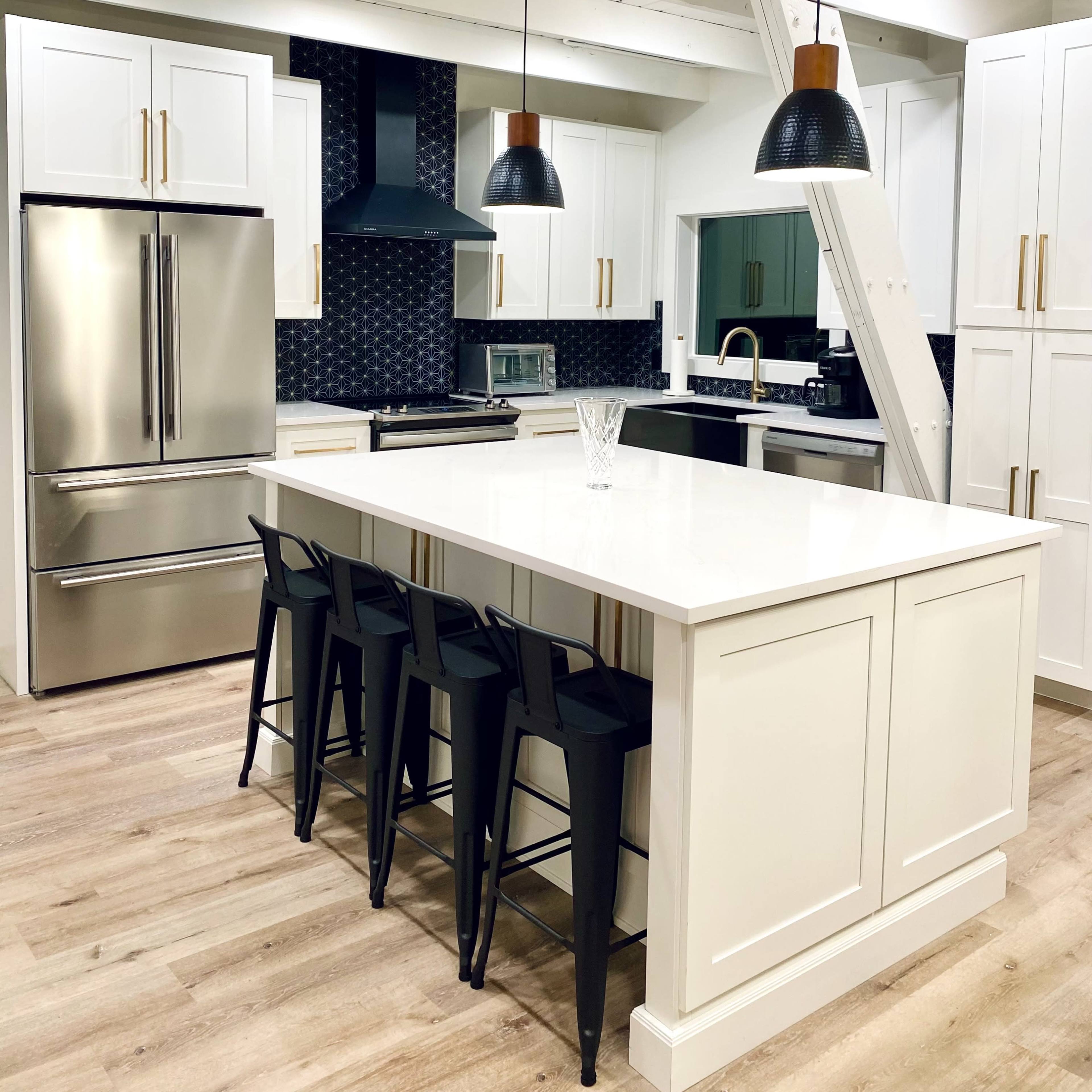 A modern kitchen with a central island, stainless steel appliances, black stools, and pendant lights hanging above.