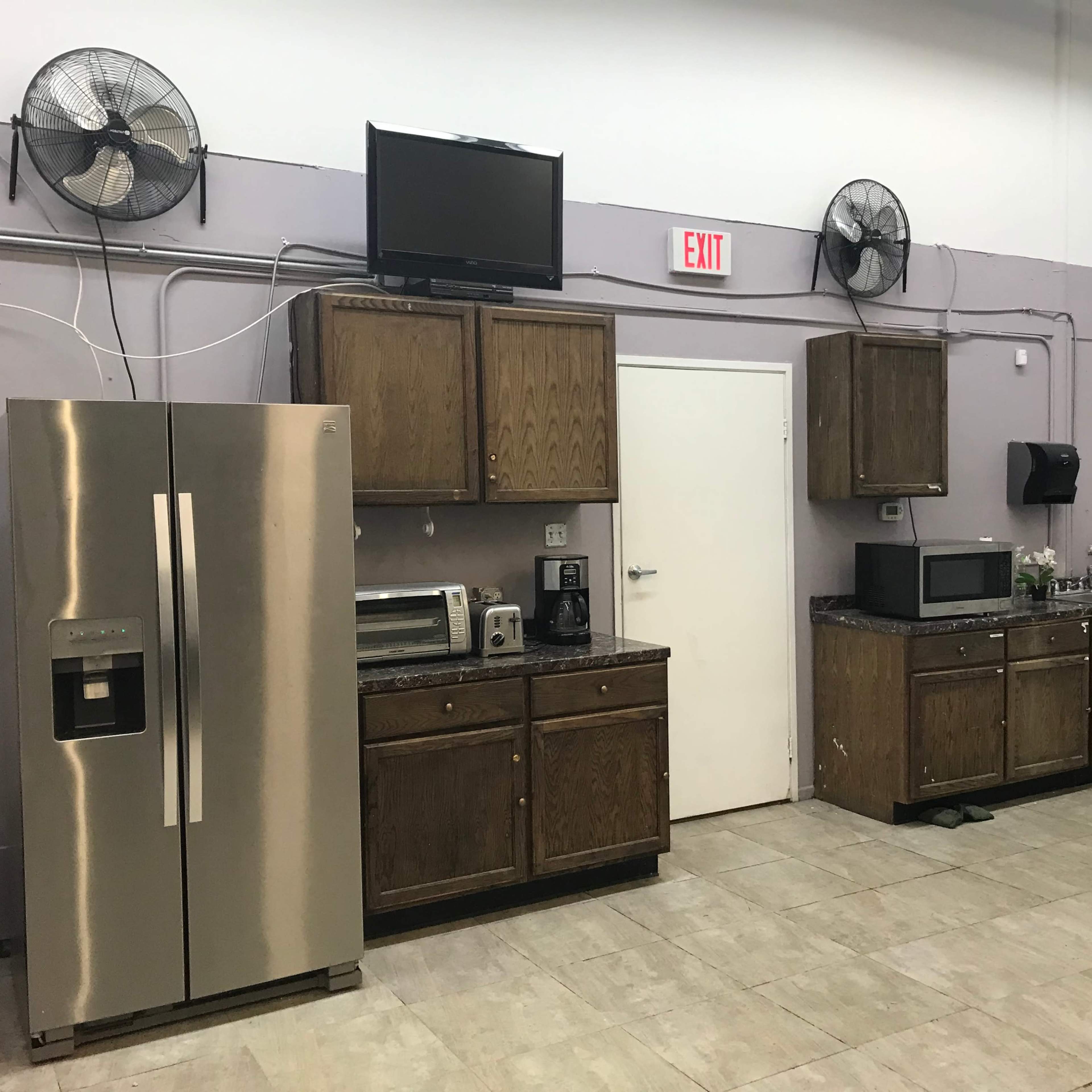 A kitchen with stainless steel appliances, including a refrigerator, microwave, and coffee maker, against a wall with wooden cabinets and ceiling fans.