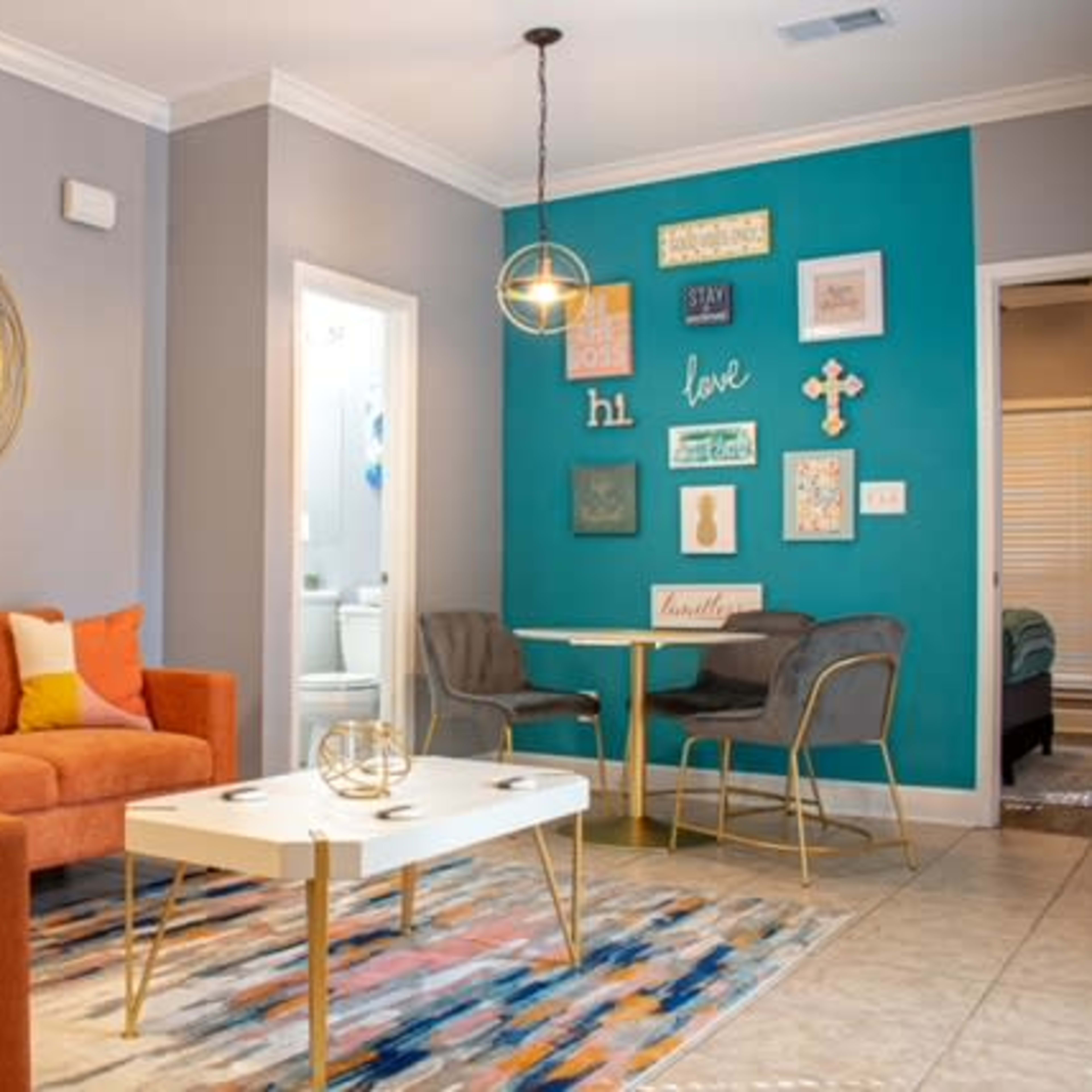 A living room with an orange couch, a round mirror, a white coffee table, and a dining area featuring teal walls adorned with framed decorations.