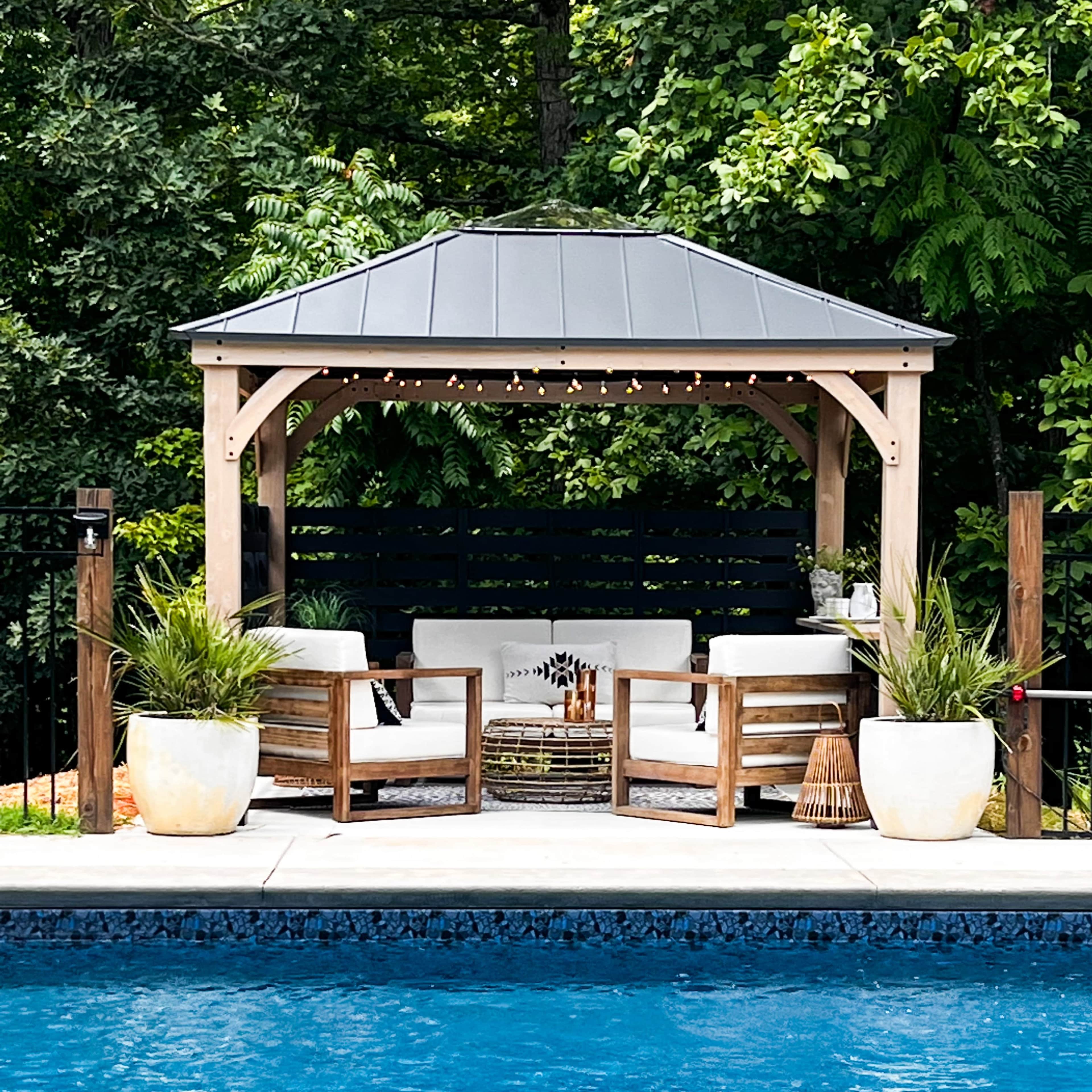 A wooden gazebo with seating is positioned beside a swimming pool, surrounded by lush greenery.