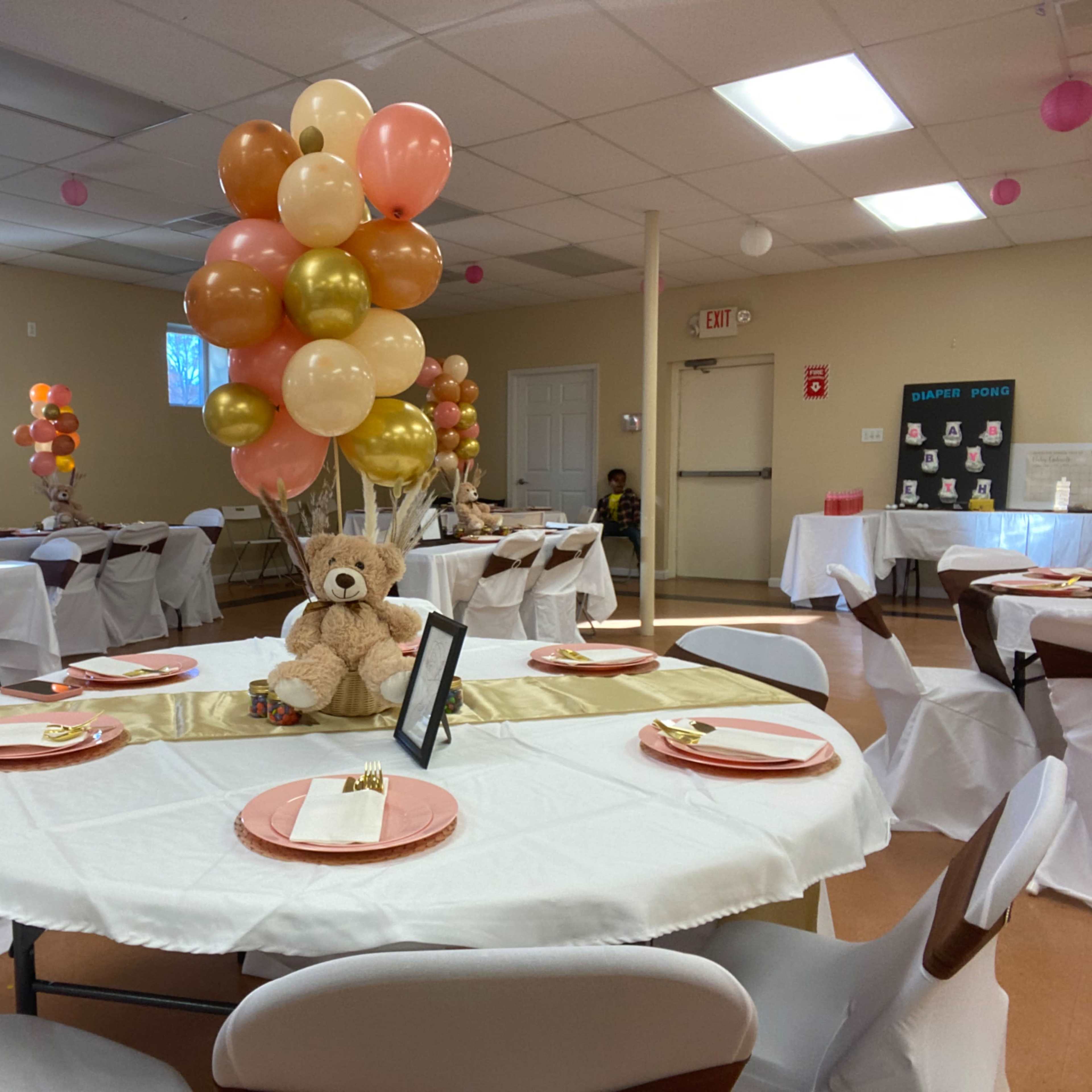 The scene shows a decorated party venue with tables set with white tablecloths, colorful balloon centerpieces, and a teddy bear at the center of each table.