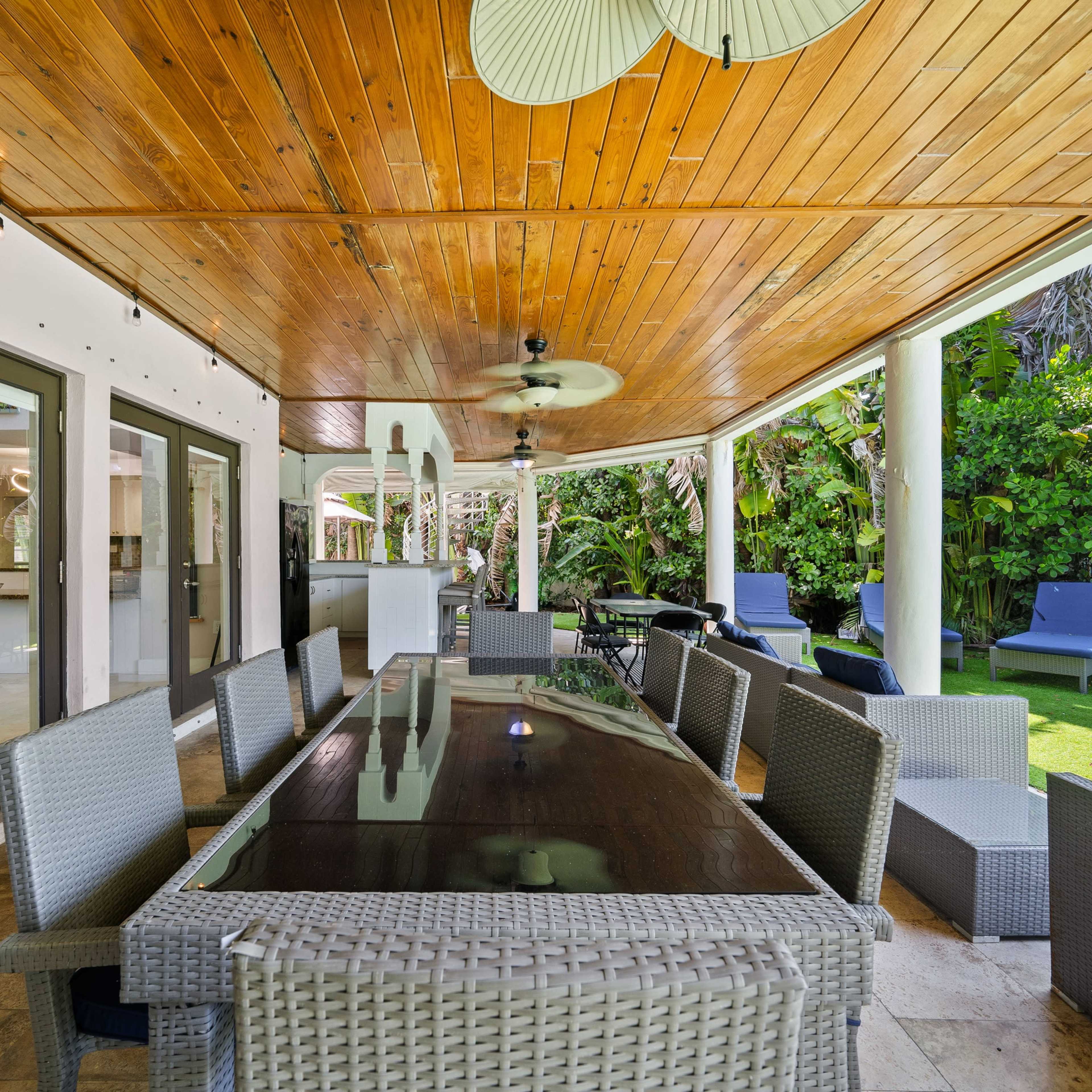 A covered outdoor dining area features a long table surrounded by wicker chairs, with a ceiling fan above and lush greenery visible in the background.