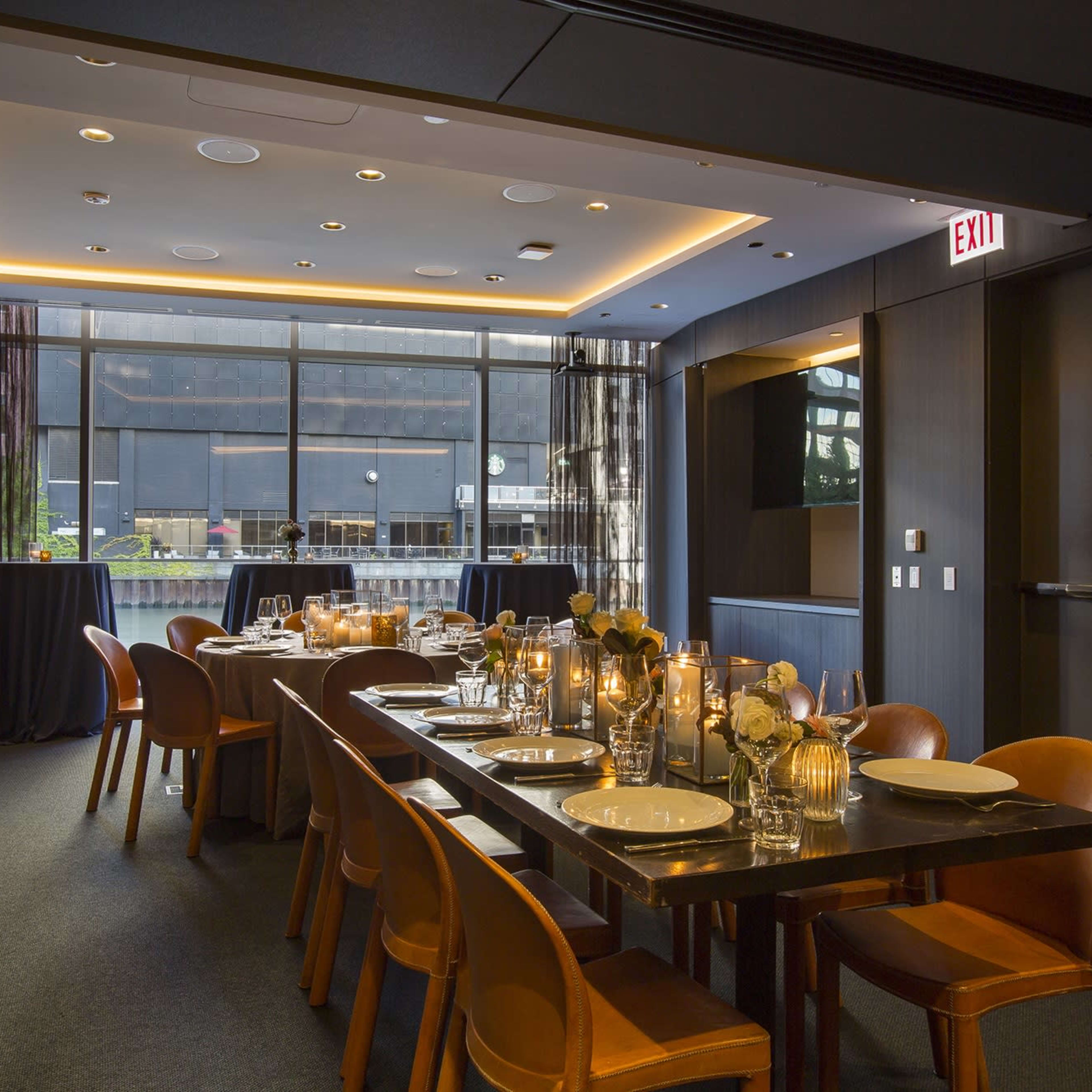 A dining room features a long table set with candles and glassware, illuminated by soft lighting and a large window displaying an outdoor view.