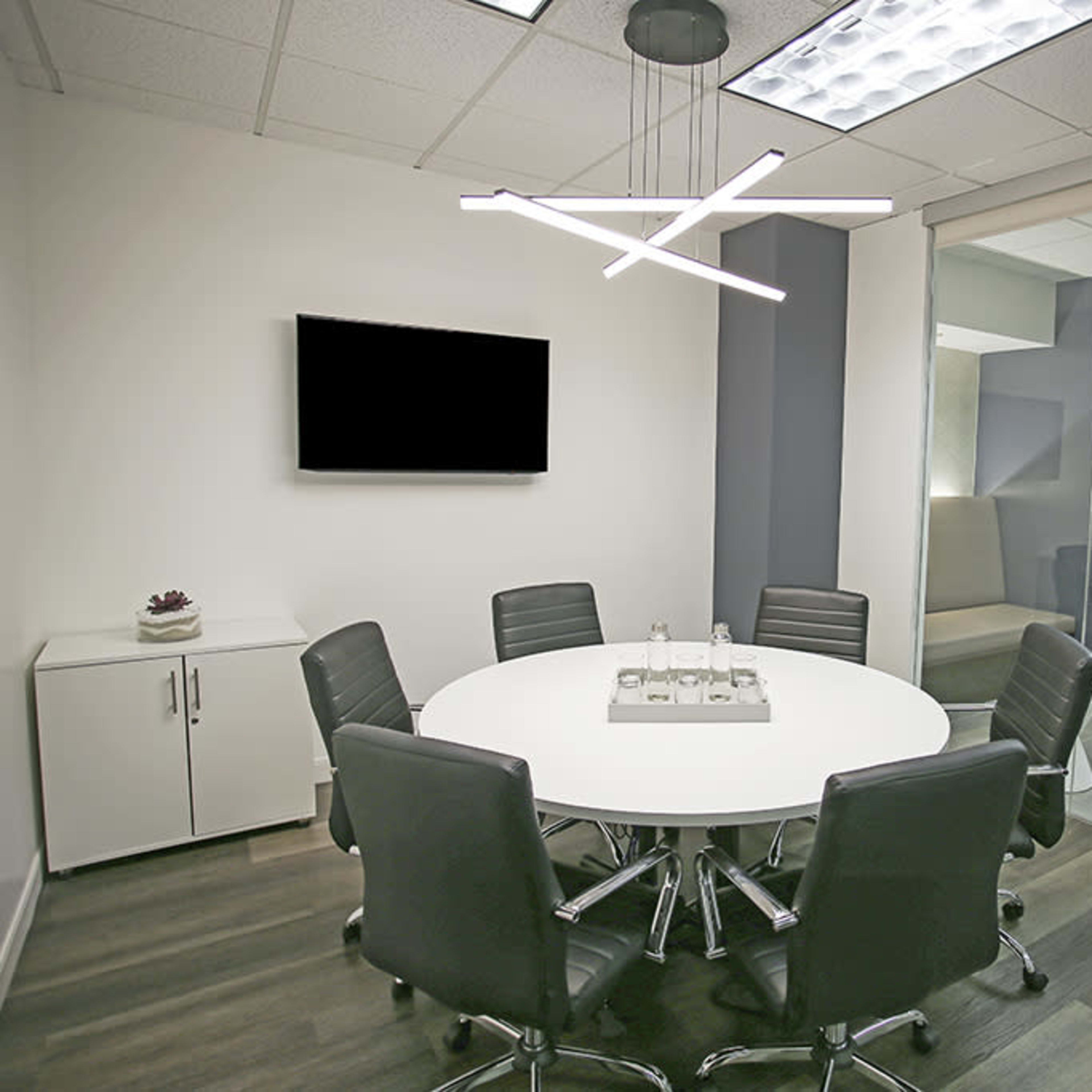 A modern conference room features a round table surrounded by black chairs, a flat-screen television on the wall, and large windows with plants visible outside.