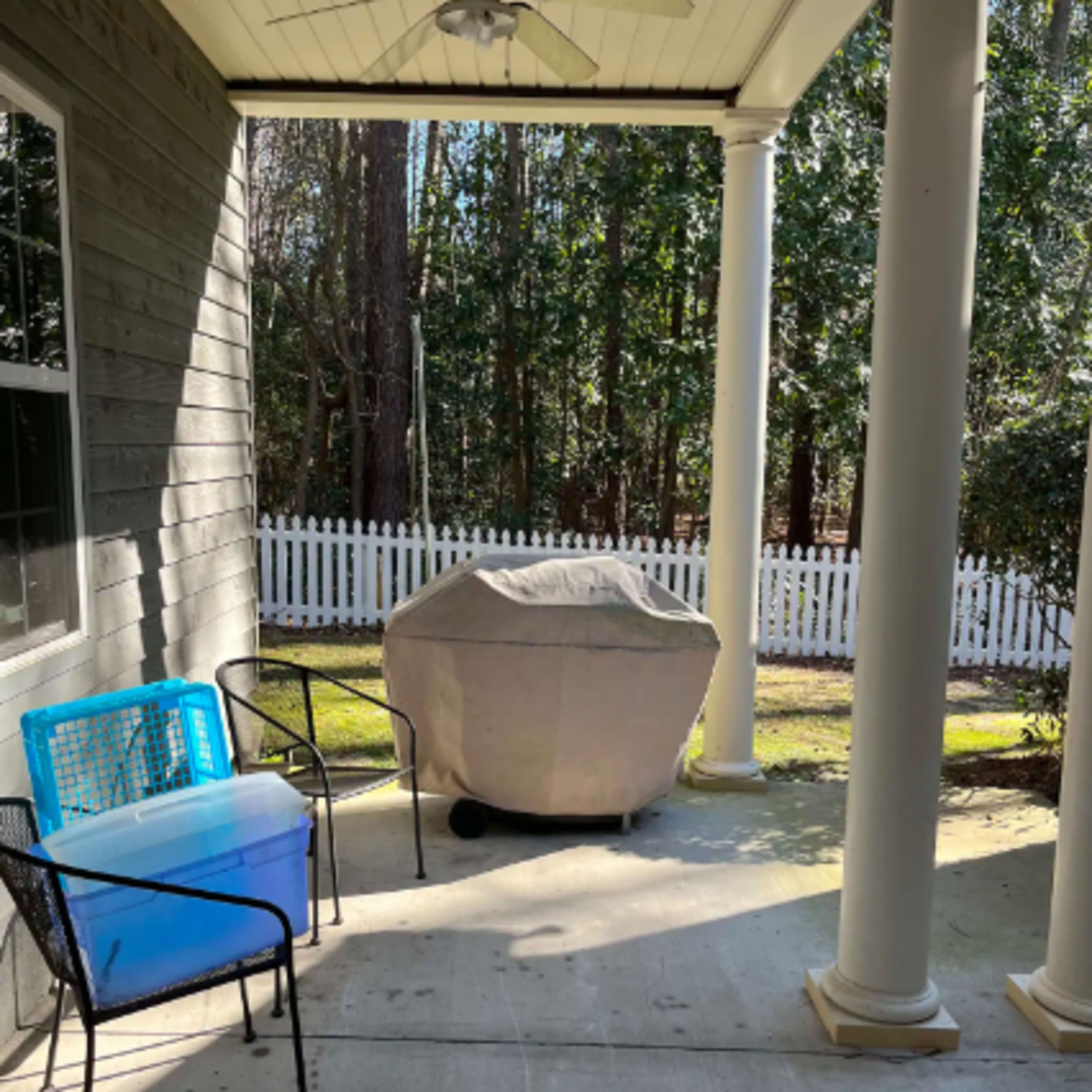 A covered grill sits on a concrete patio, flanked by two chairs and surrounded by trees and a white picket fence.