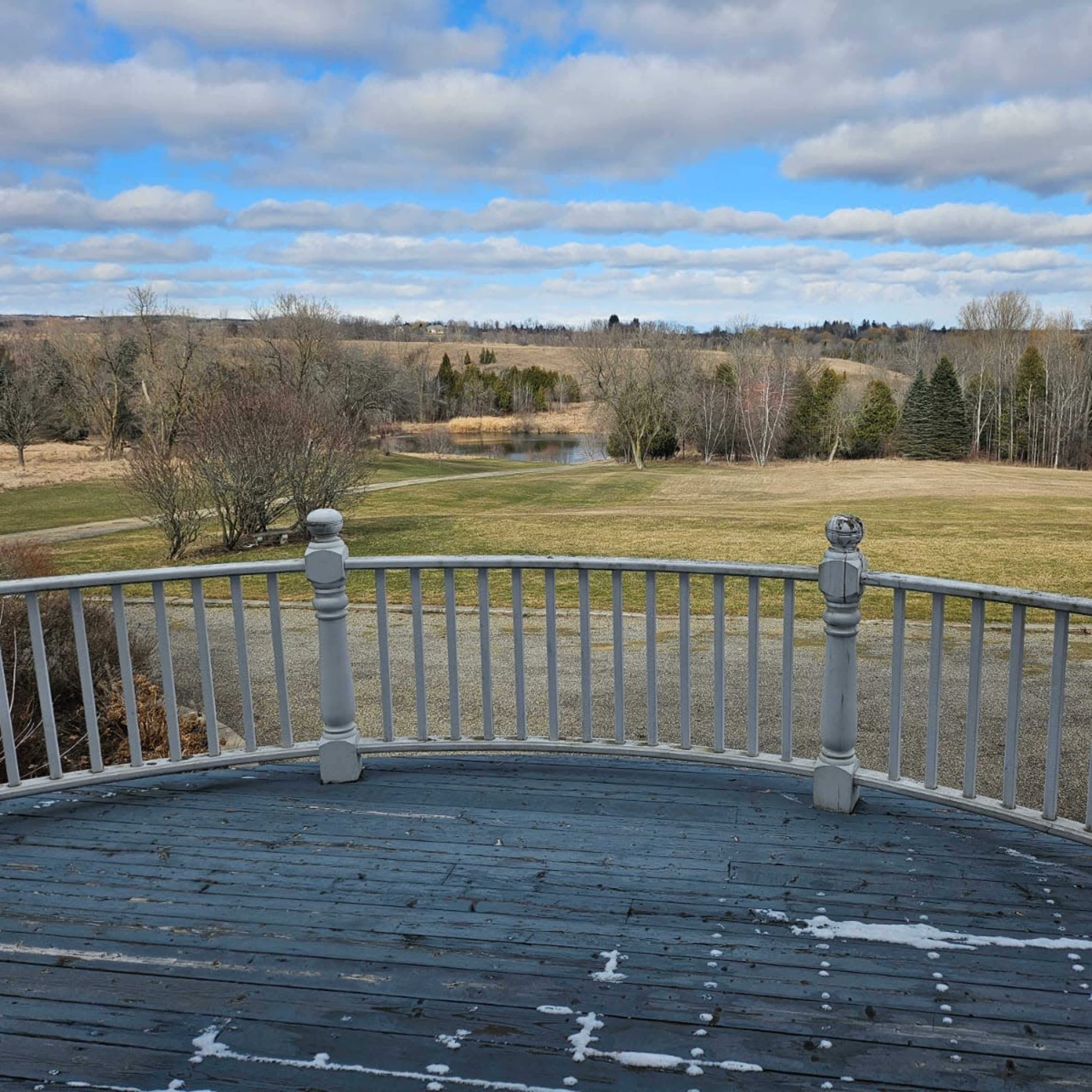 A balcony view overlooking a grassy field with sparse trees and a small body of water in the distance under a partly cloudy sky.