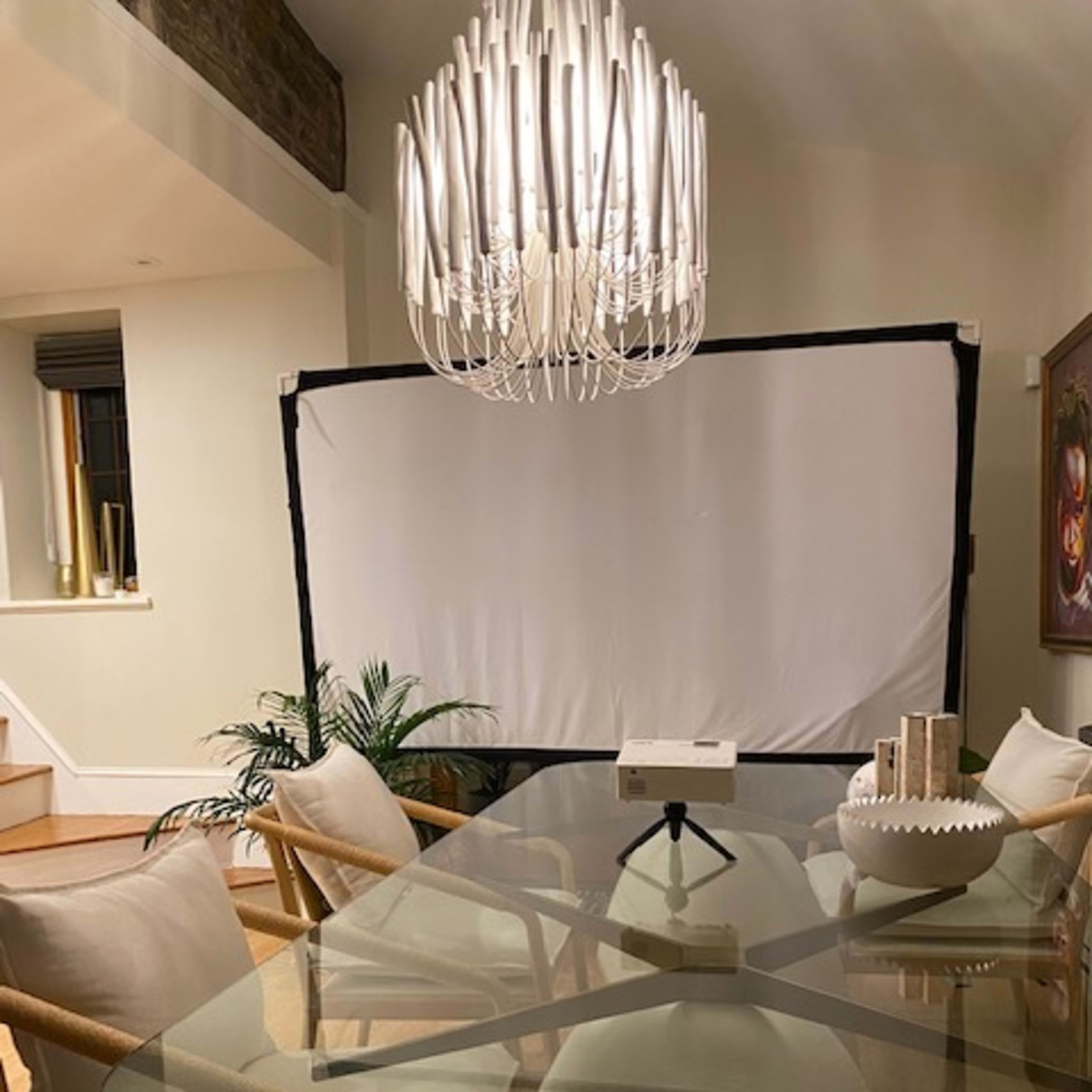 A dining area features a glass table surrounded by beige chairs, with a large white backdrop set up in the background and a modern chandelier overhead.