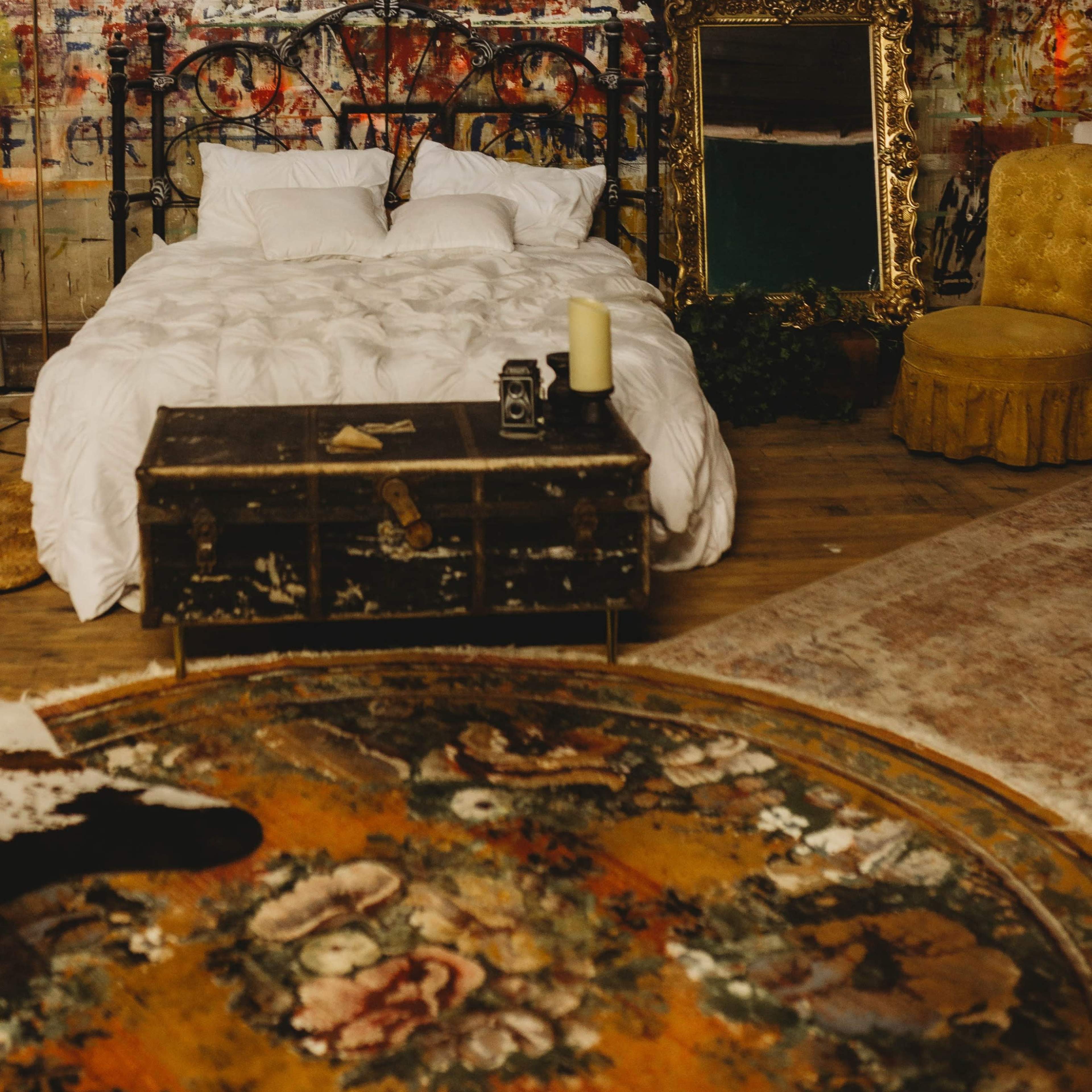 A bedroom features a wrought-iron bed with white bedding, a vintage chandelier, a distressed brick wall covered in colorful paint, and a gold-framed mirror beside a decorative yellow chair.
