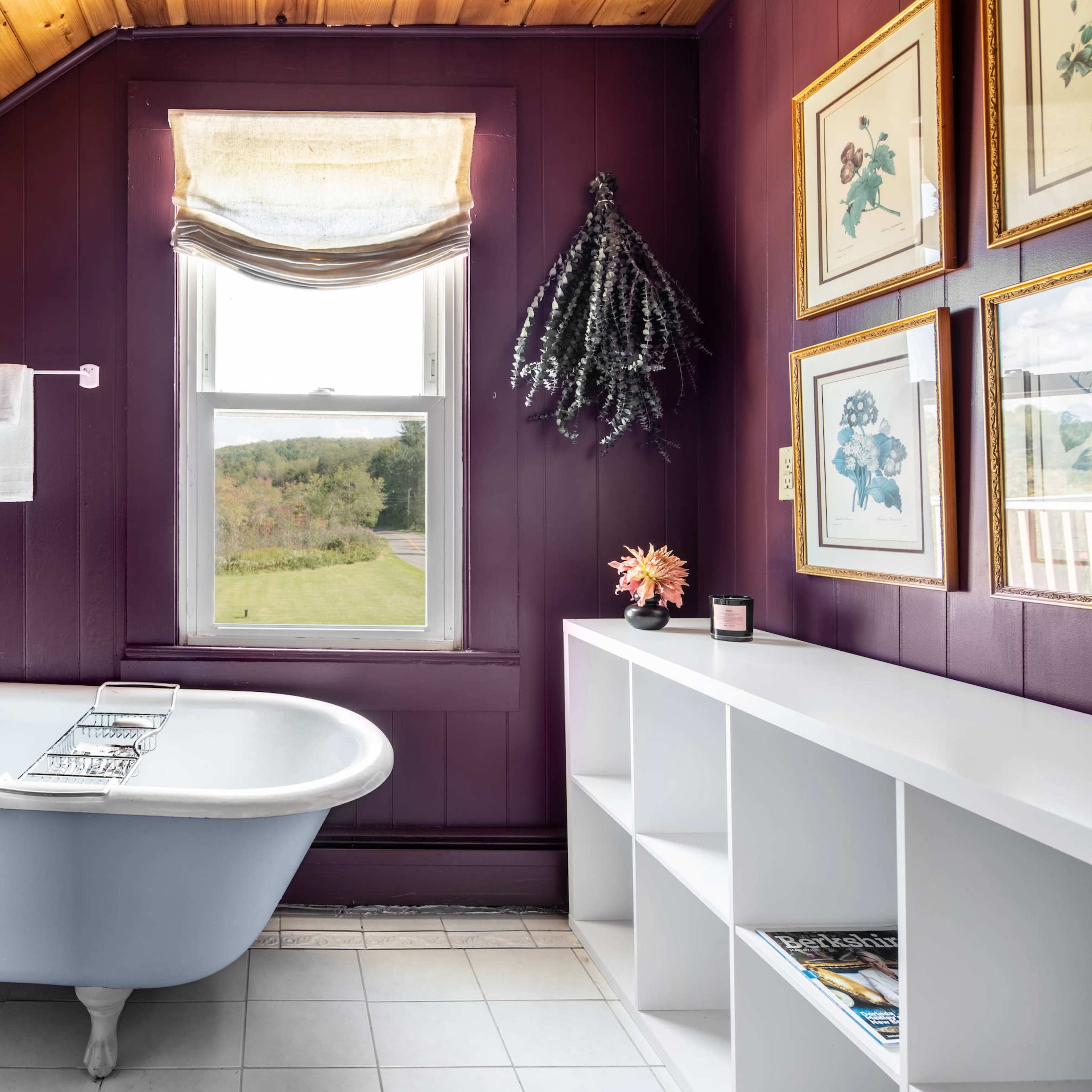A bathroom with a vintage clawfoot bathtub, purple walls, a window providing natural light, and a small shelf with decor.