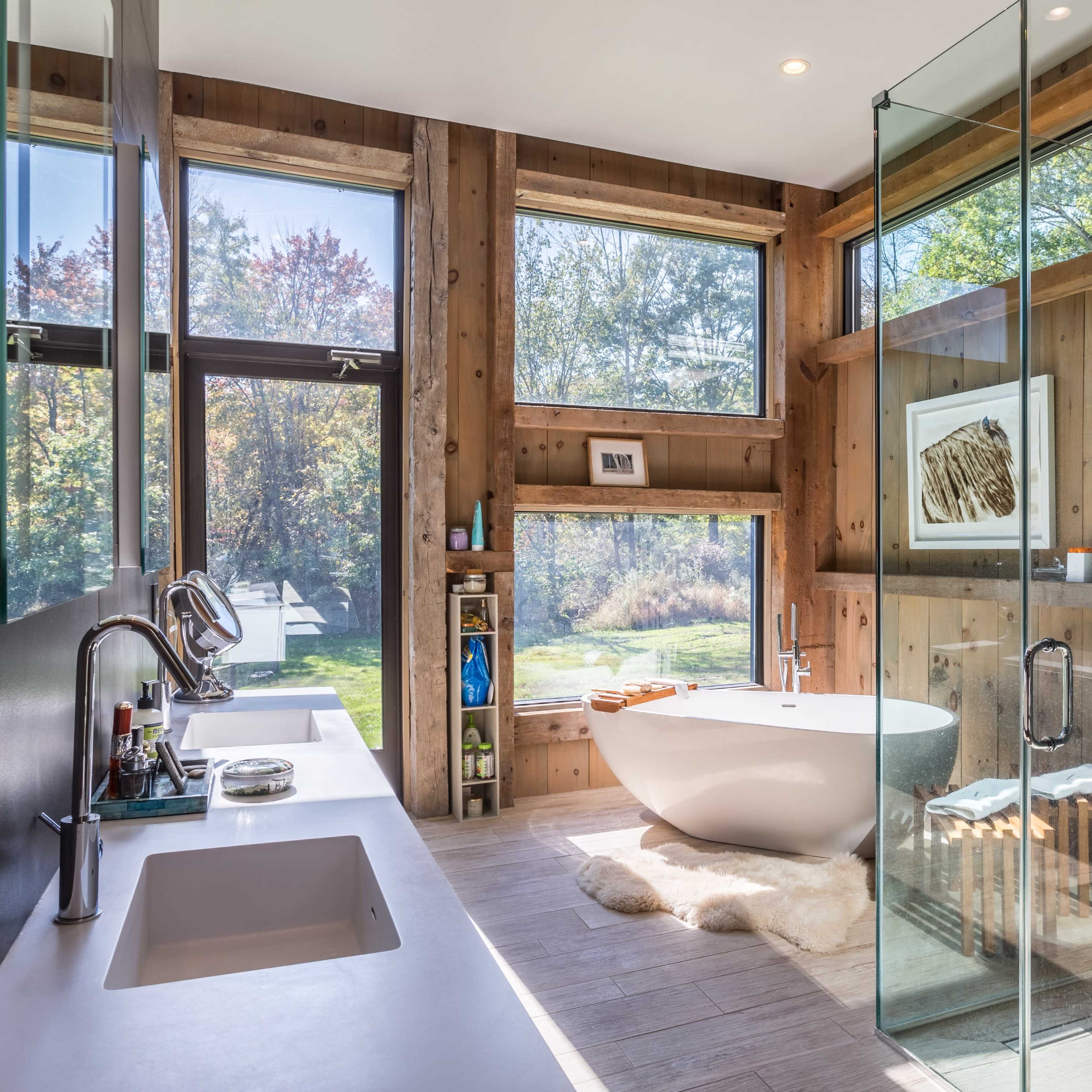 A modern bathroom features a freestanding bathtub by large windows, with wooden walls and a glass shower enclosure.