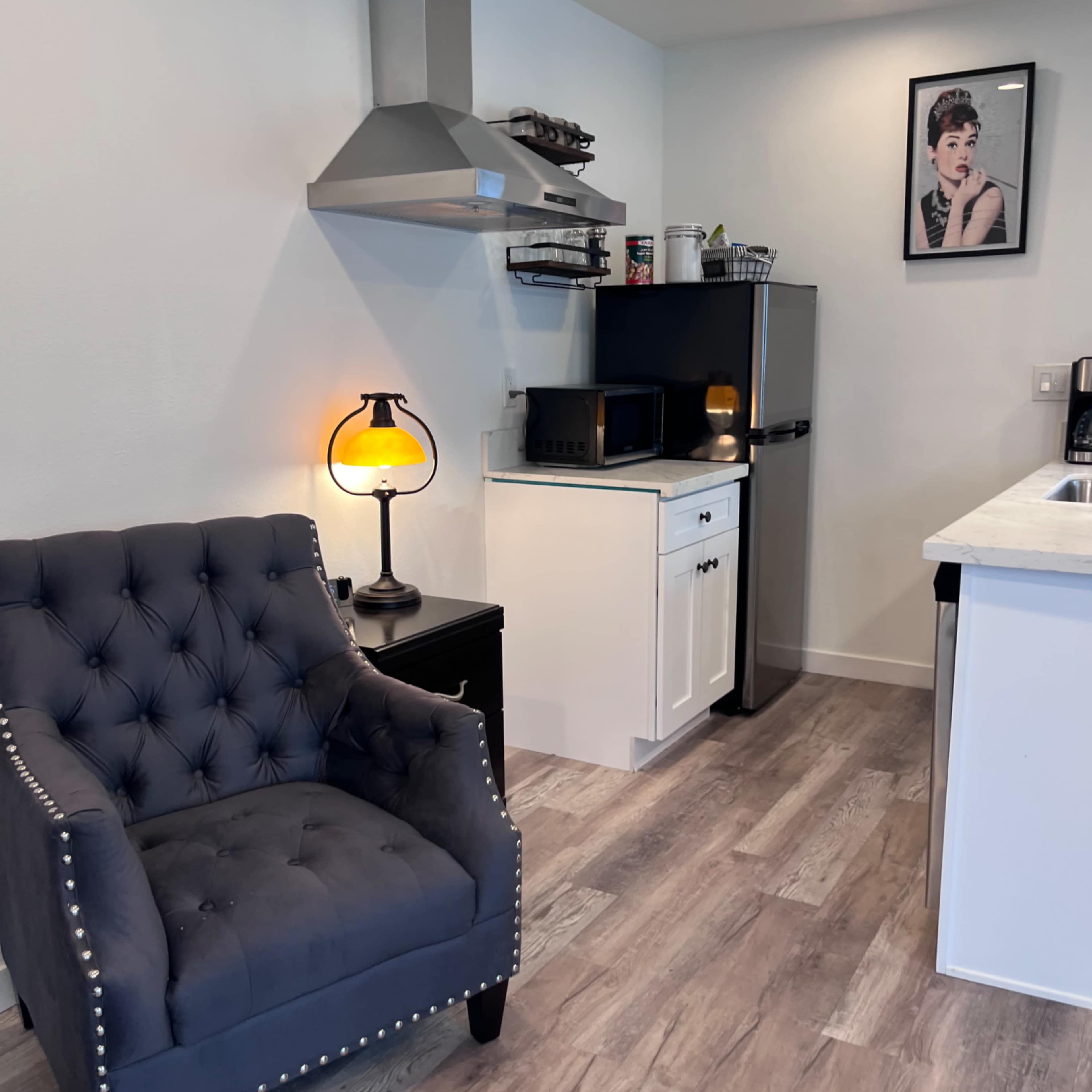 A modern kitchen features a tufted armchair, a stainless steel range hood, a white countertop, and a black refrigerator.