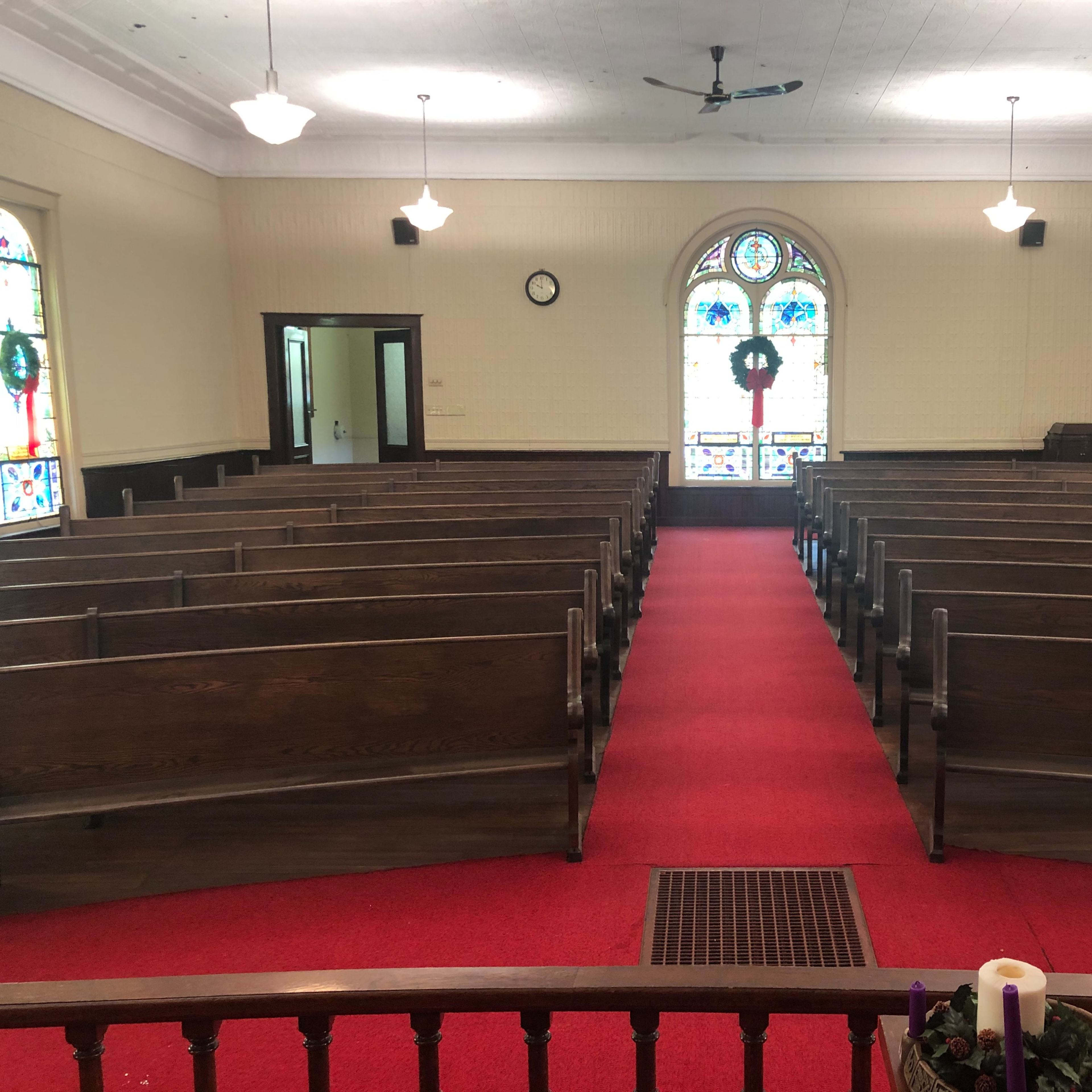 A church interior with wooden pews arranged in rows on red carpet, facing a stained glass window at the front.