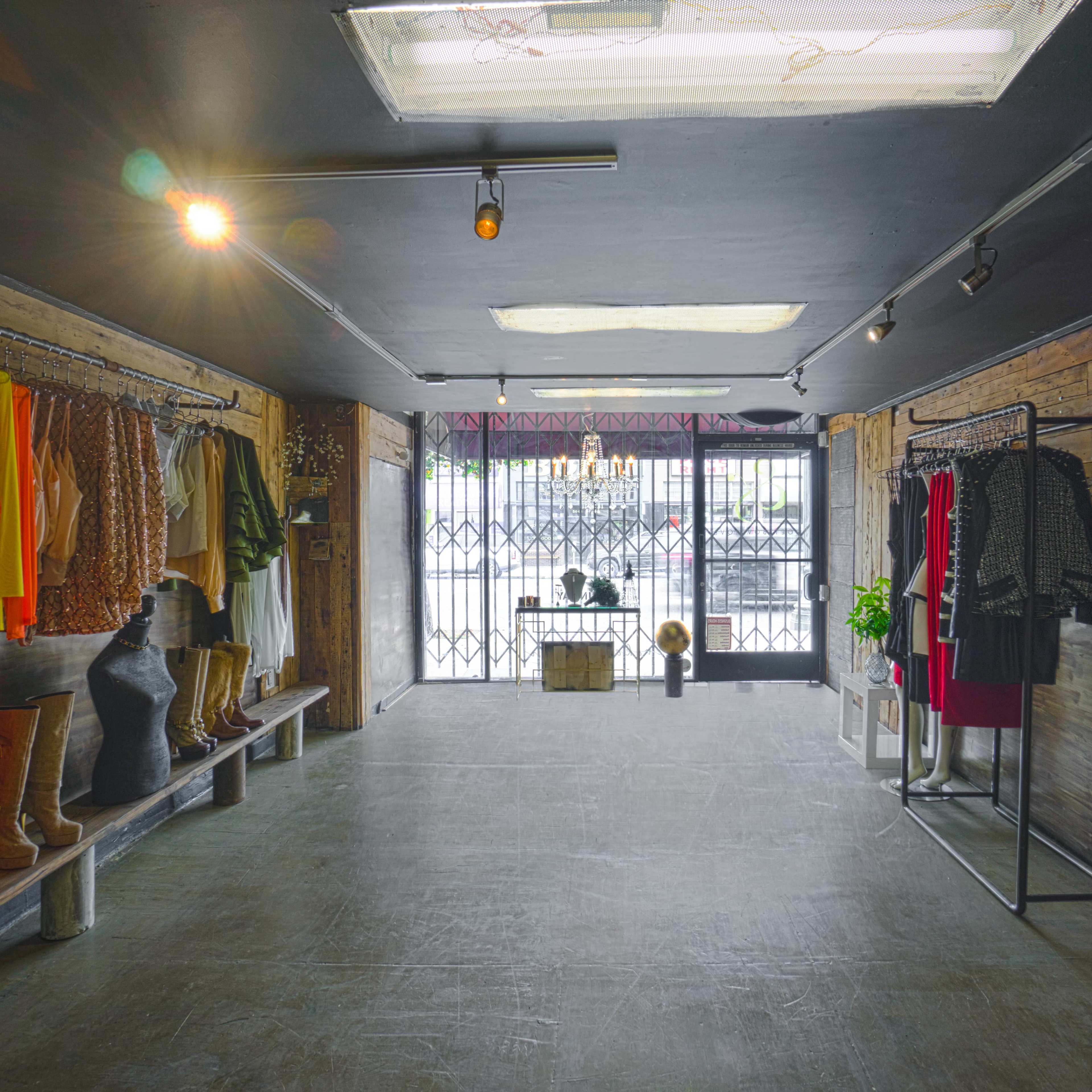 The interior of a clothing boutique features clothing racks with various garments, wooden accents, and a large window with barred security.