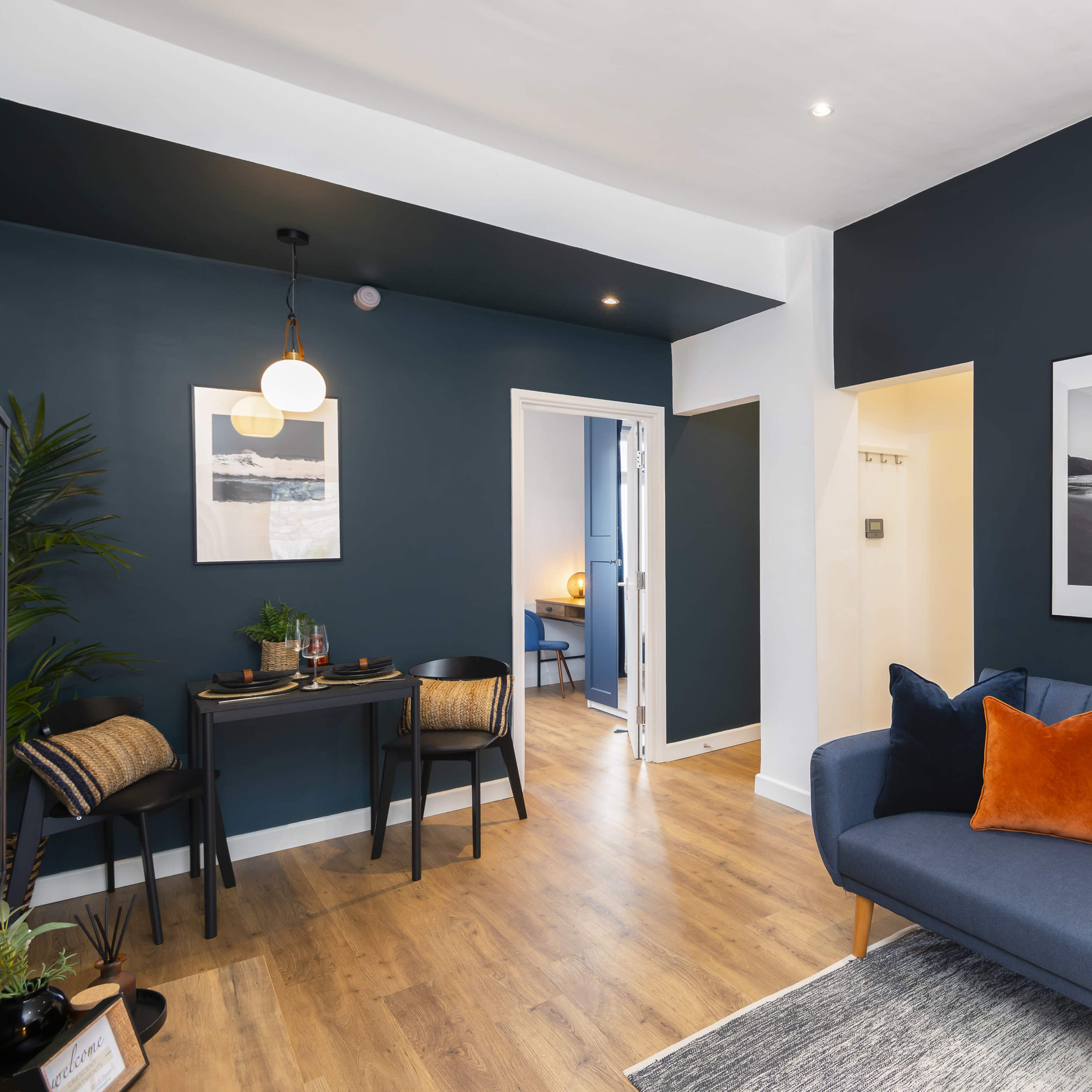 A modern living space featuring a blue couch with orange pillows, a dining area with a table and two chairs, and framed photographs on the dark blue wall.