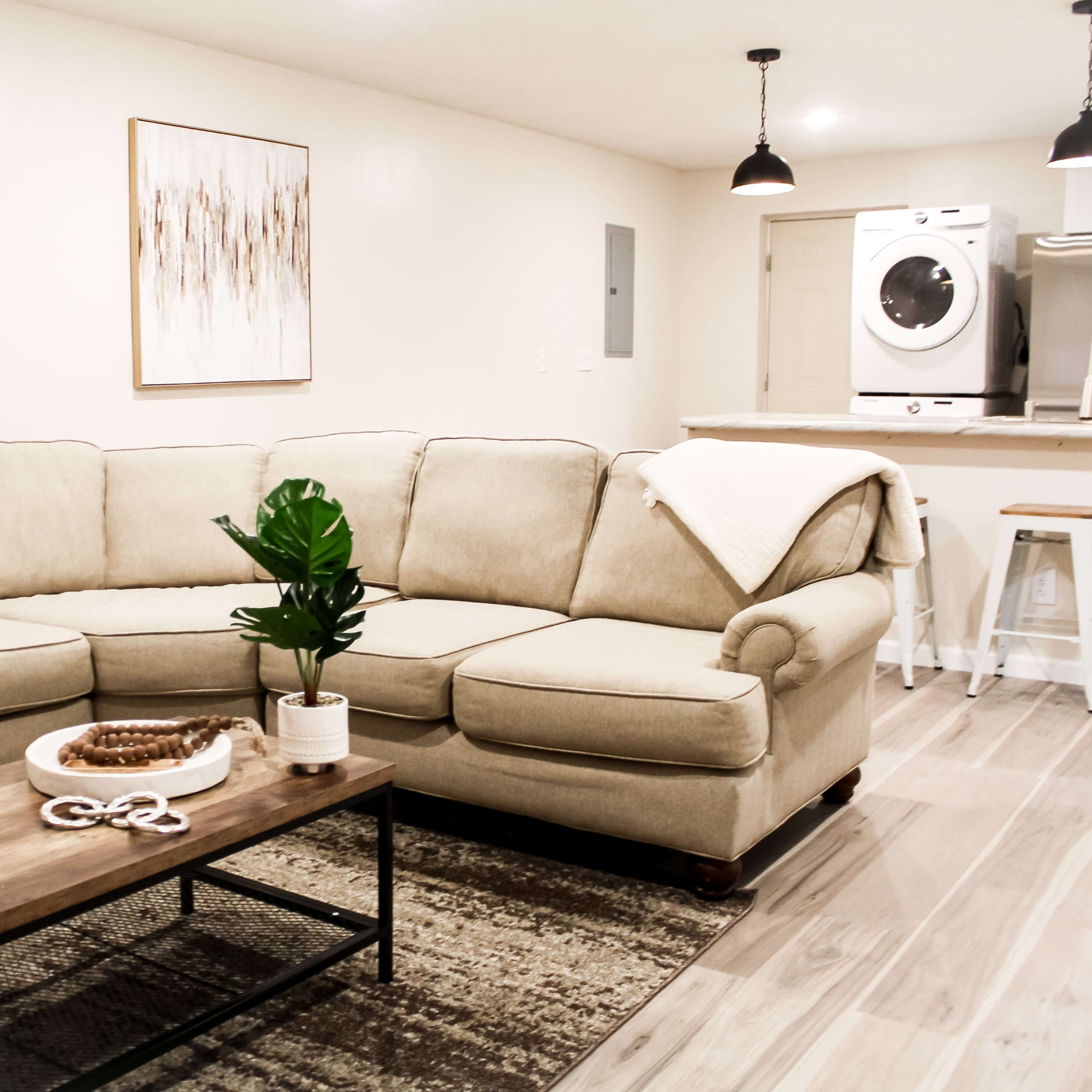 A beige sectional sofa is positioned in a living room with a wooden coffee table, a small potted plant, and a laundry area visible in the background.