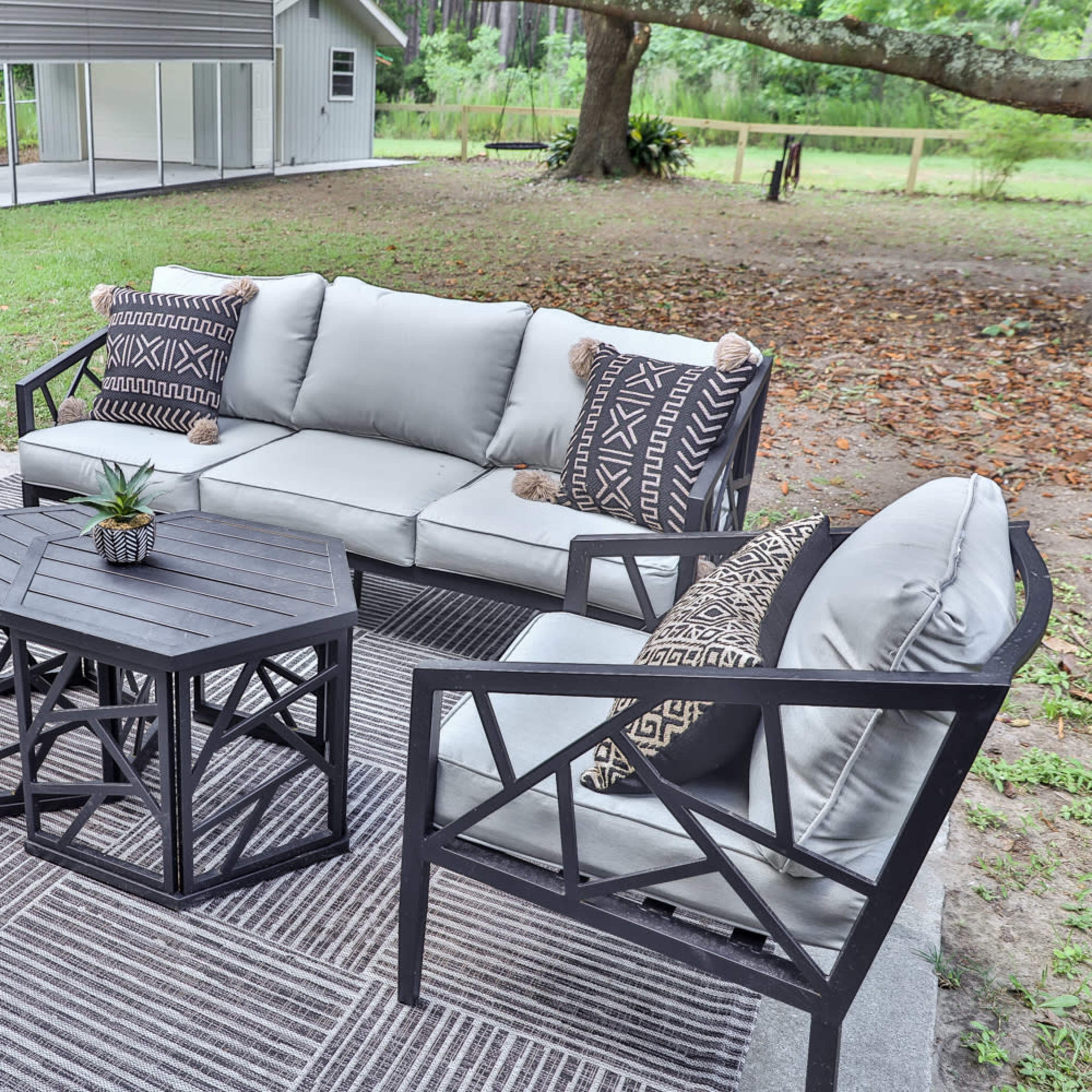 A modern outdoor seating area features a gray sofa and chairs with patterned pillows, arranged around a black, geometric coffee table on a grassy lawn.