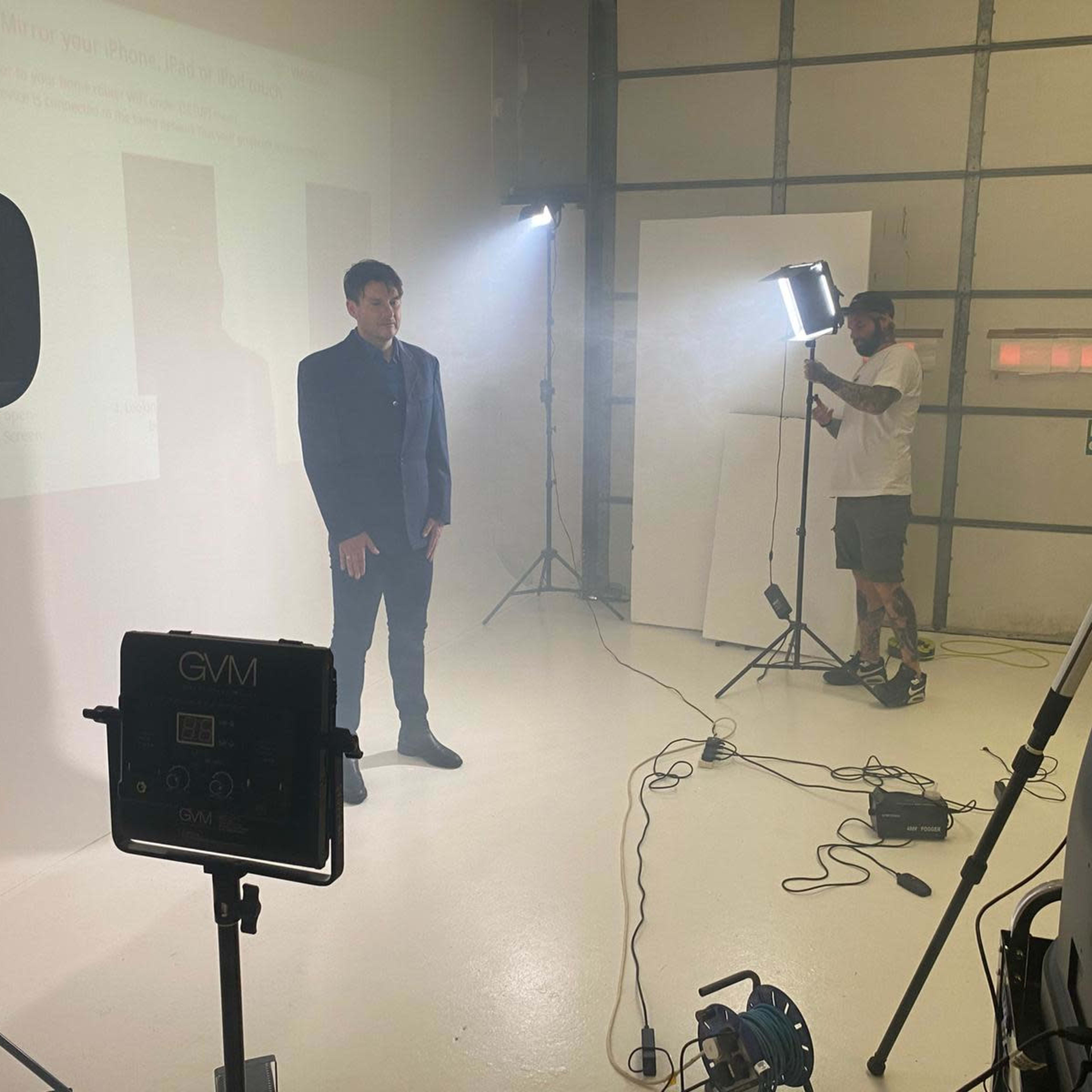 A man in a suit stands in a studio surrounded by lighting equipment and a camera operator.