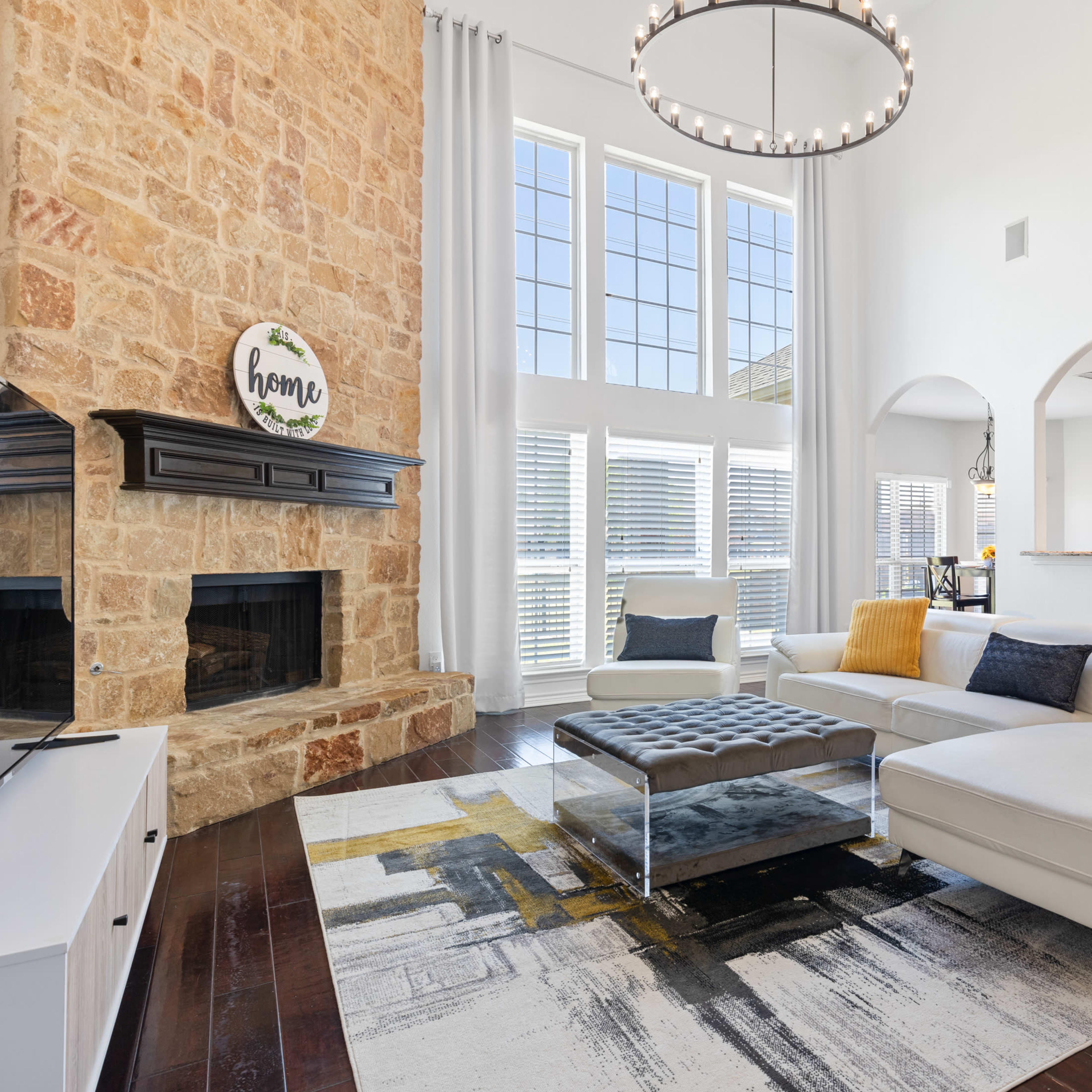 A modern living room with a stone fireplace, large windows, and a light-colored sectional sofa positioned on a patterned area rug.