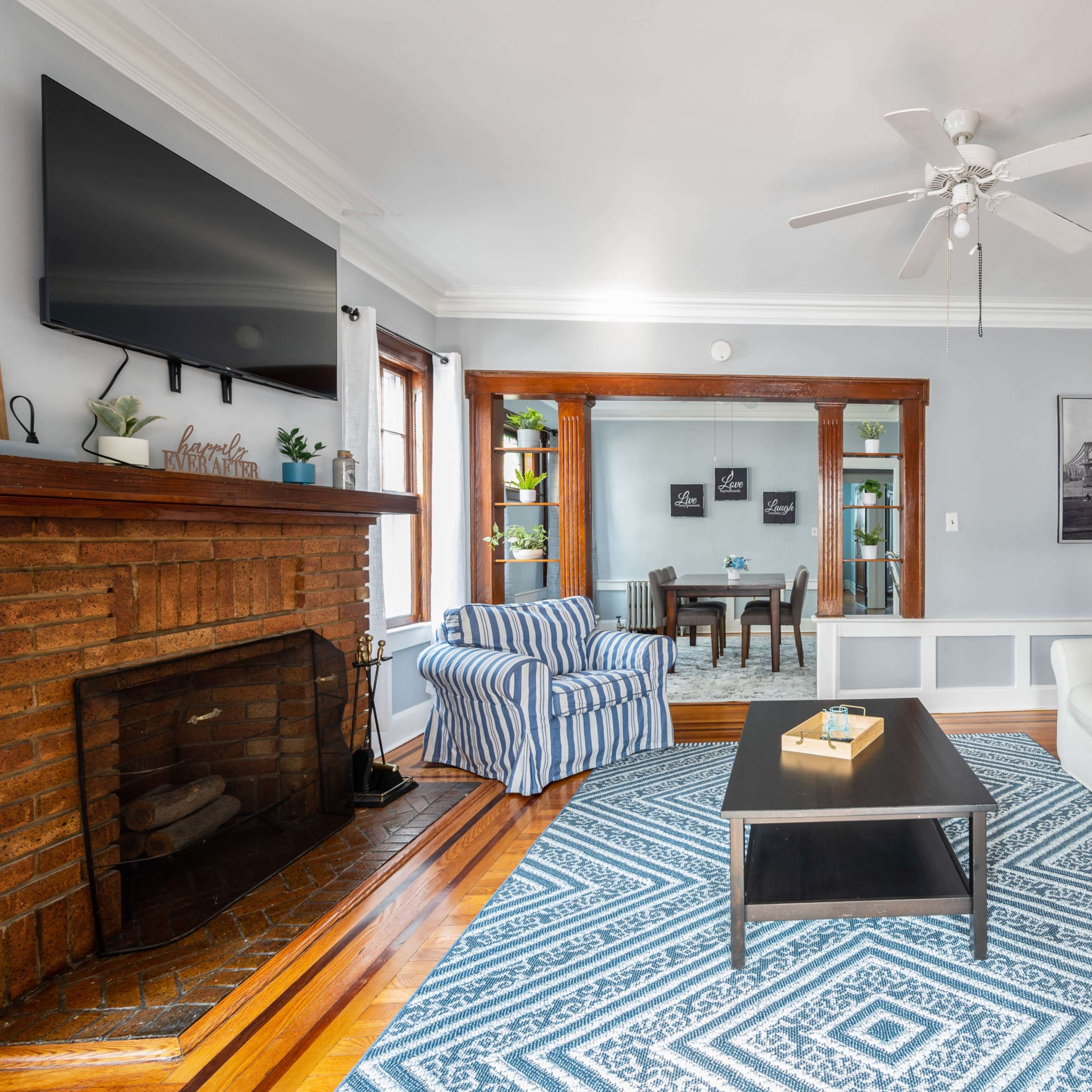 The living room features a white sofa, a striped blue chair, a coffee table, and a fireplace, with a television mounted on the wall and art displayed around the room.