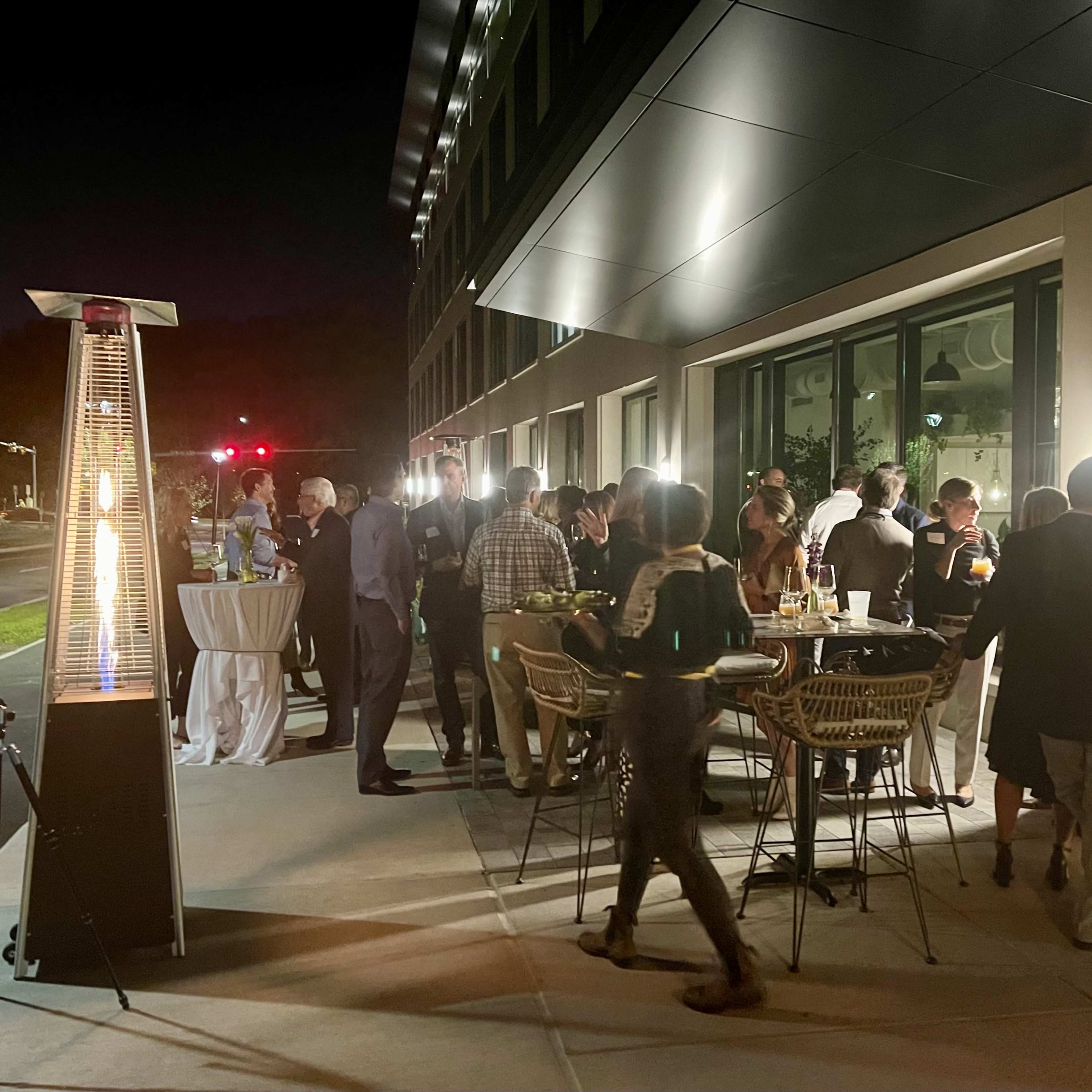 A group of people socialize at an outdoor gathering next to a modern building, illuminated by gas heaters and ambient lighting.