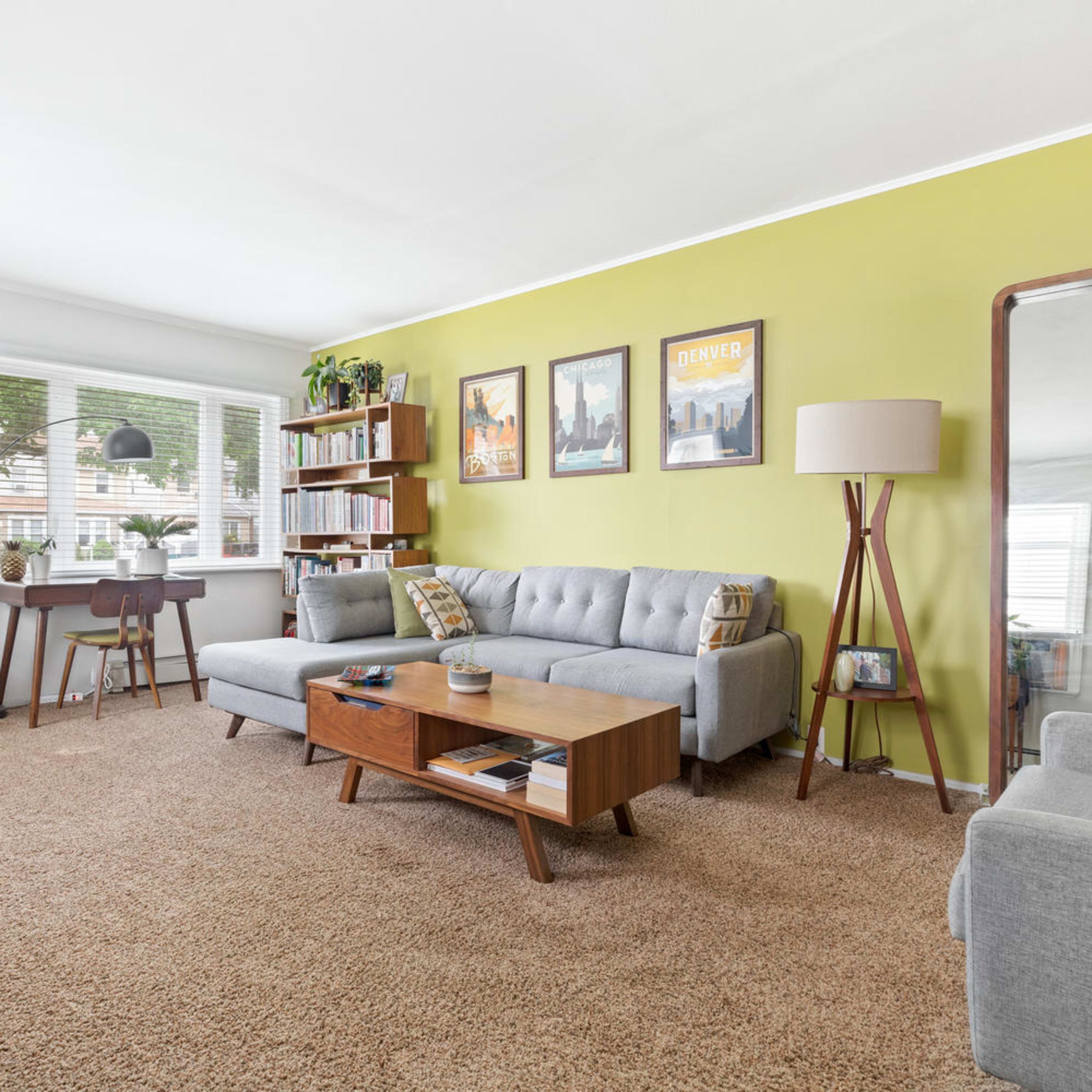 A cozy living room features a gray sofa, a wooden coffee table, and a small workspace by a window, with green walls and various decorative plants.