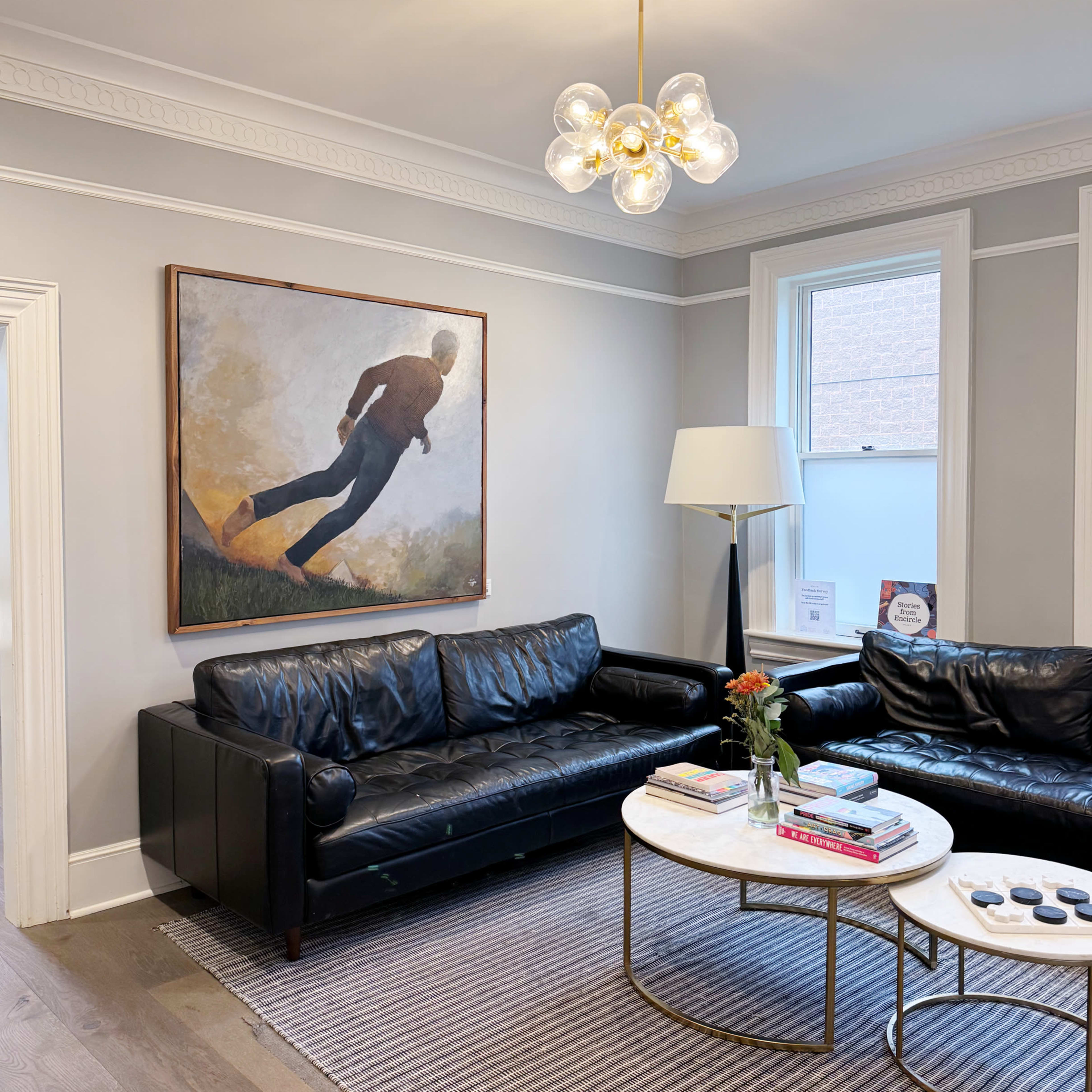 The living room features two black leather sofas, a round coffee table with magazines, and a large artwork of a figure in motion on the wall, illuminated by a modern chandelier.