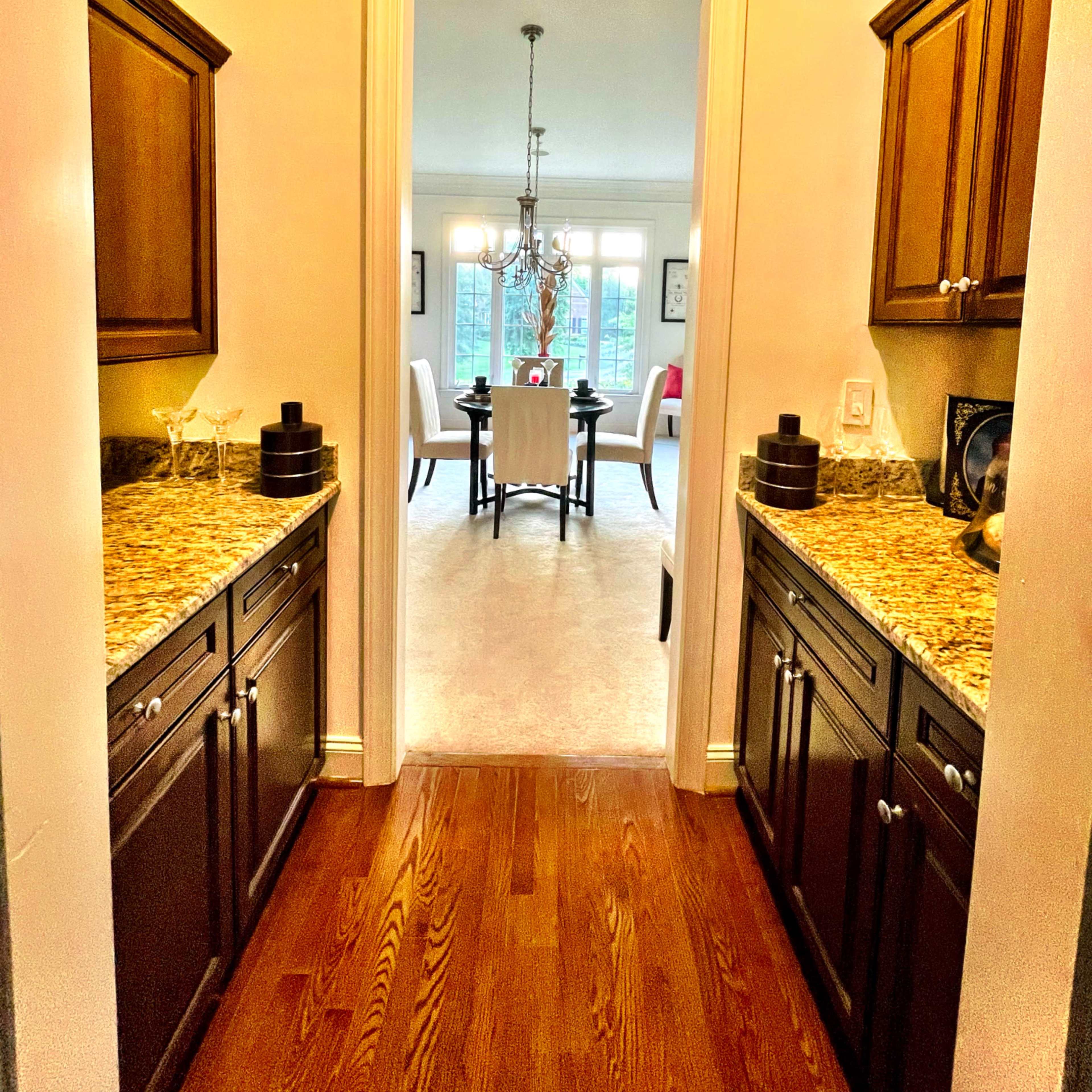 A hallway with wooden cabinetry and granite countertops leading to a dining area with a chandelier and seating.
