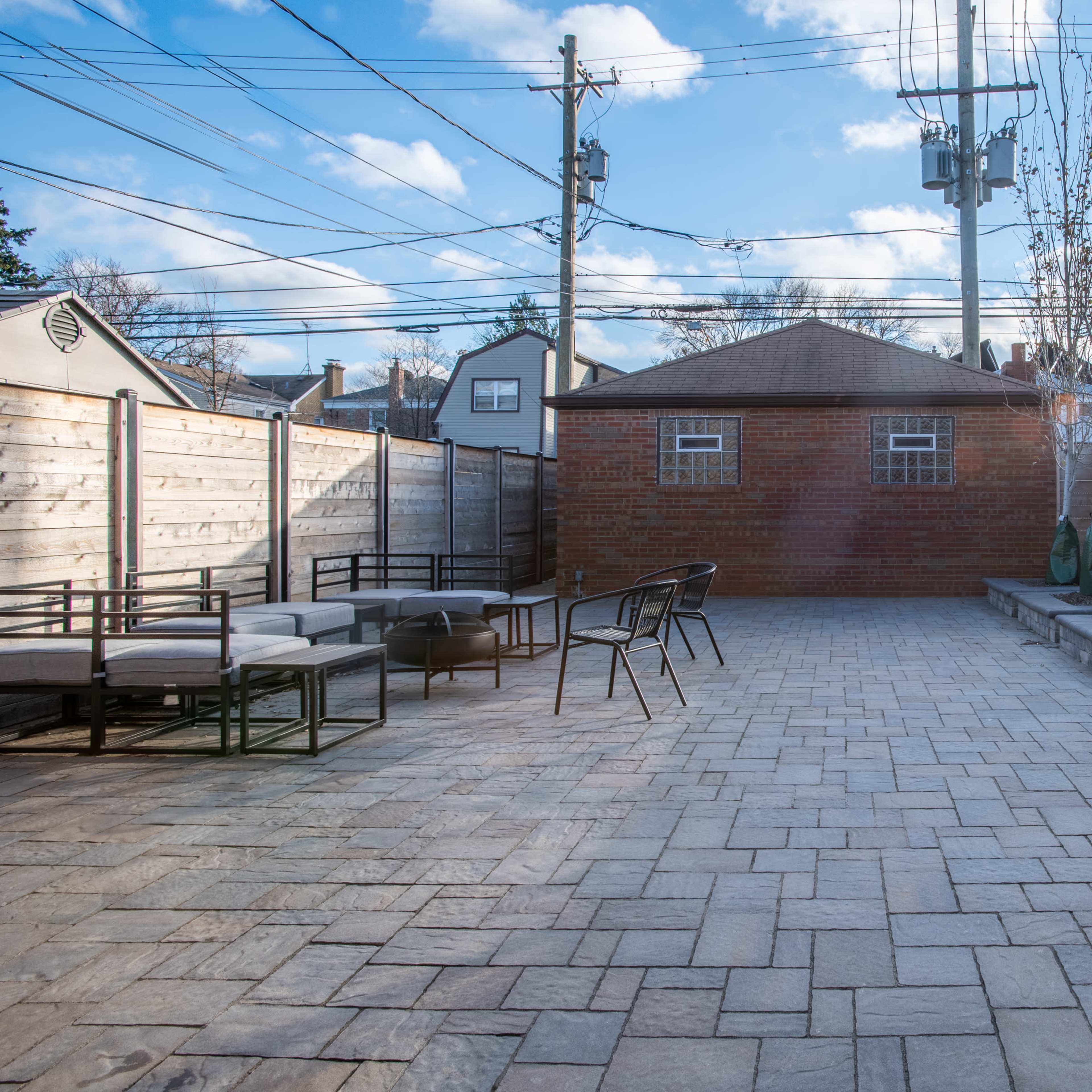 A stone-paved outdoor patio area with seating arrangements and planted trees surrounded by wooden fences and utility poles.
