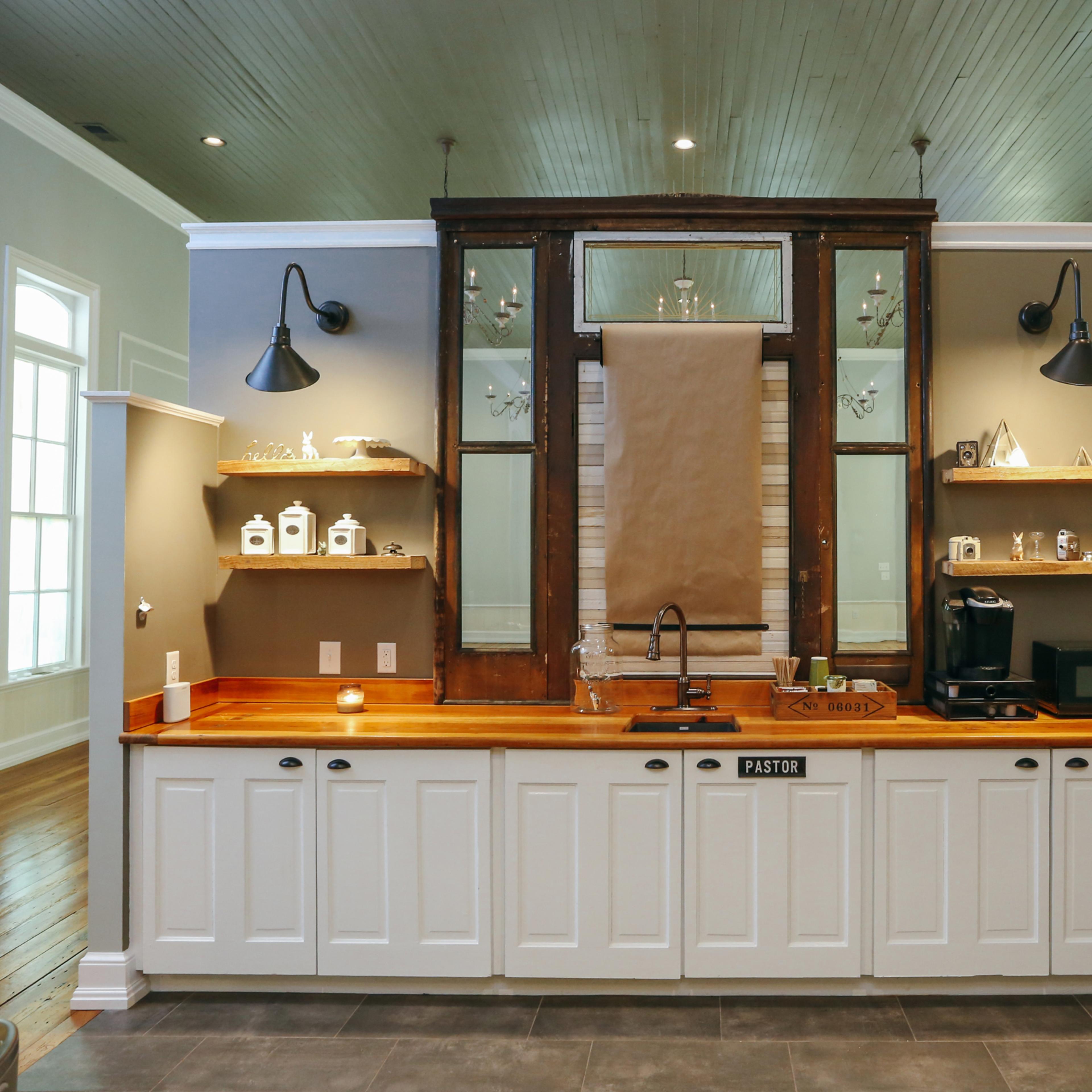 A modern kitchen with white cabinetry, a wooden countertop, and shelves displaying decorative items, set against gray walls and large windows.