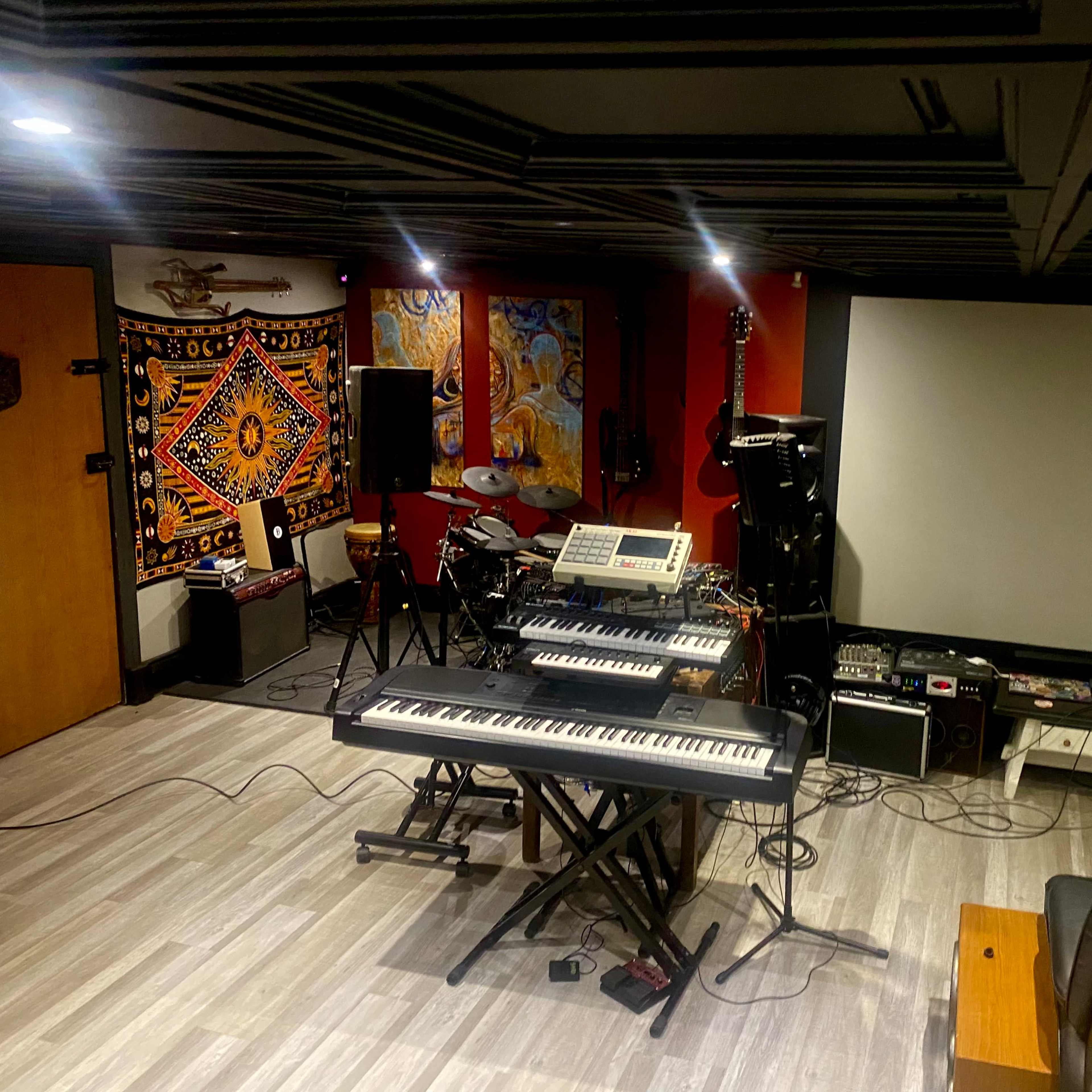 A music studio features a keyboard, electronic drum set, guitar amps, and a large screen in a dimly lit room with a colorful tapestry on one wall.