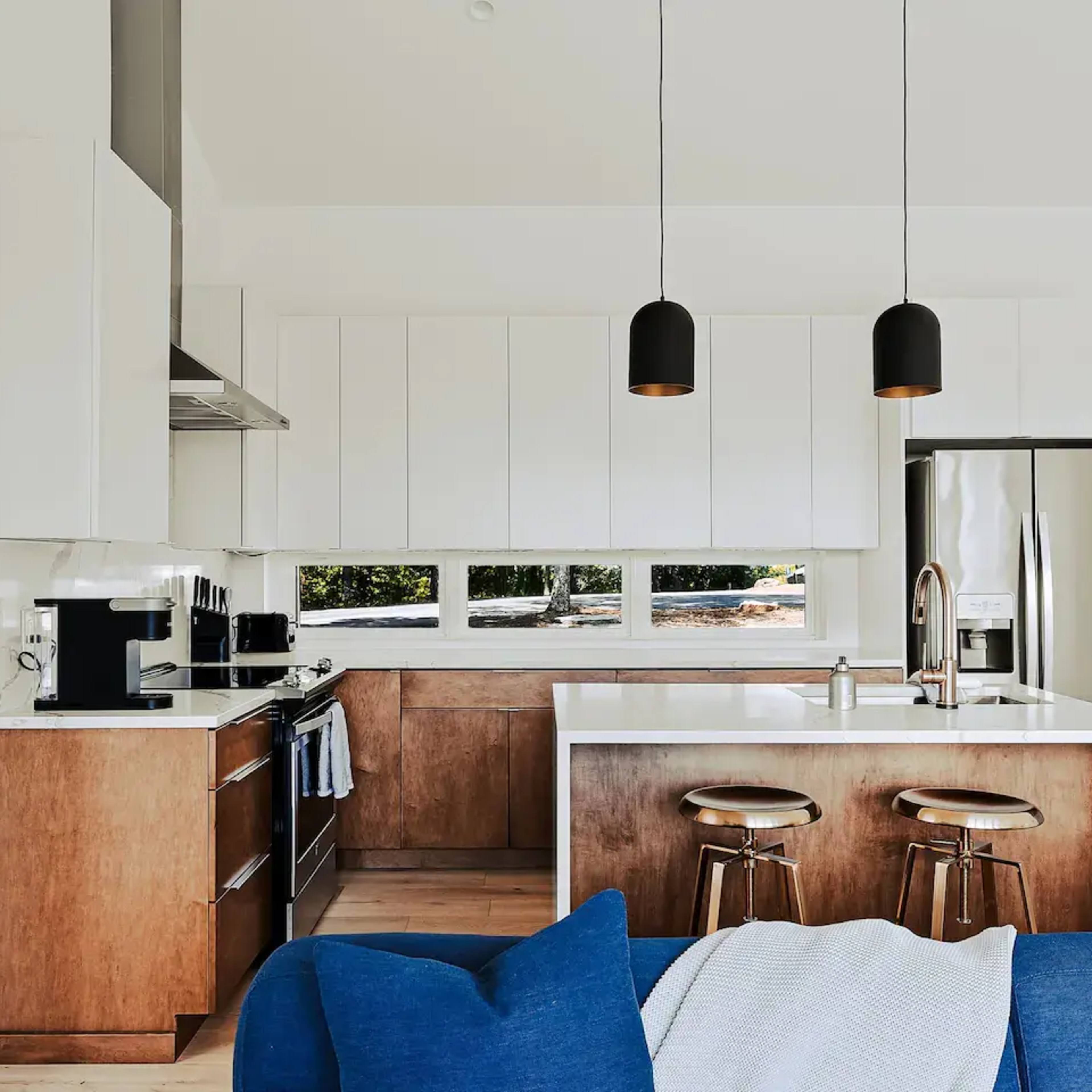 A modern kitchen features sleek cabinetry, a central island with bar stools, and large windows allowing natural light.