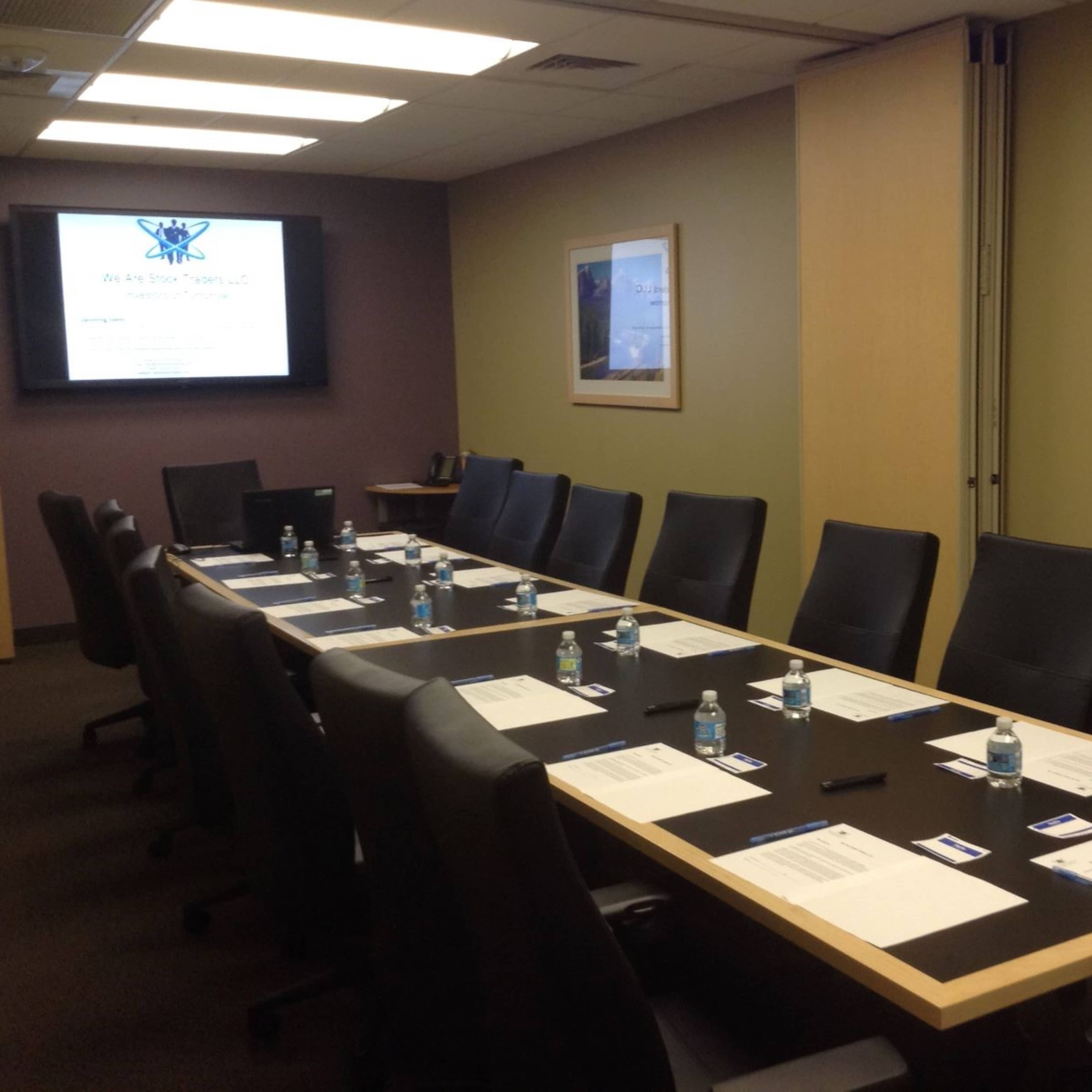 A large conference room features a long table set with water bottles and papers, with a presentation screen displayed at one end.