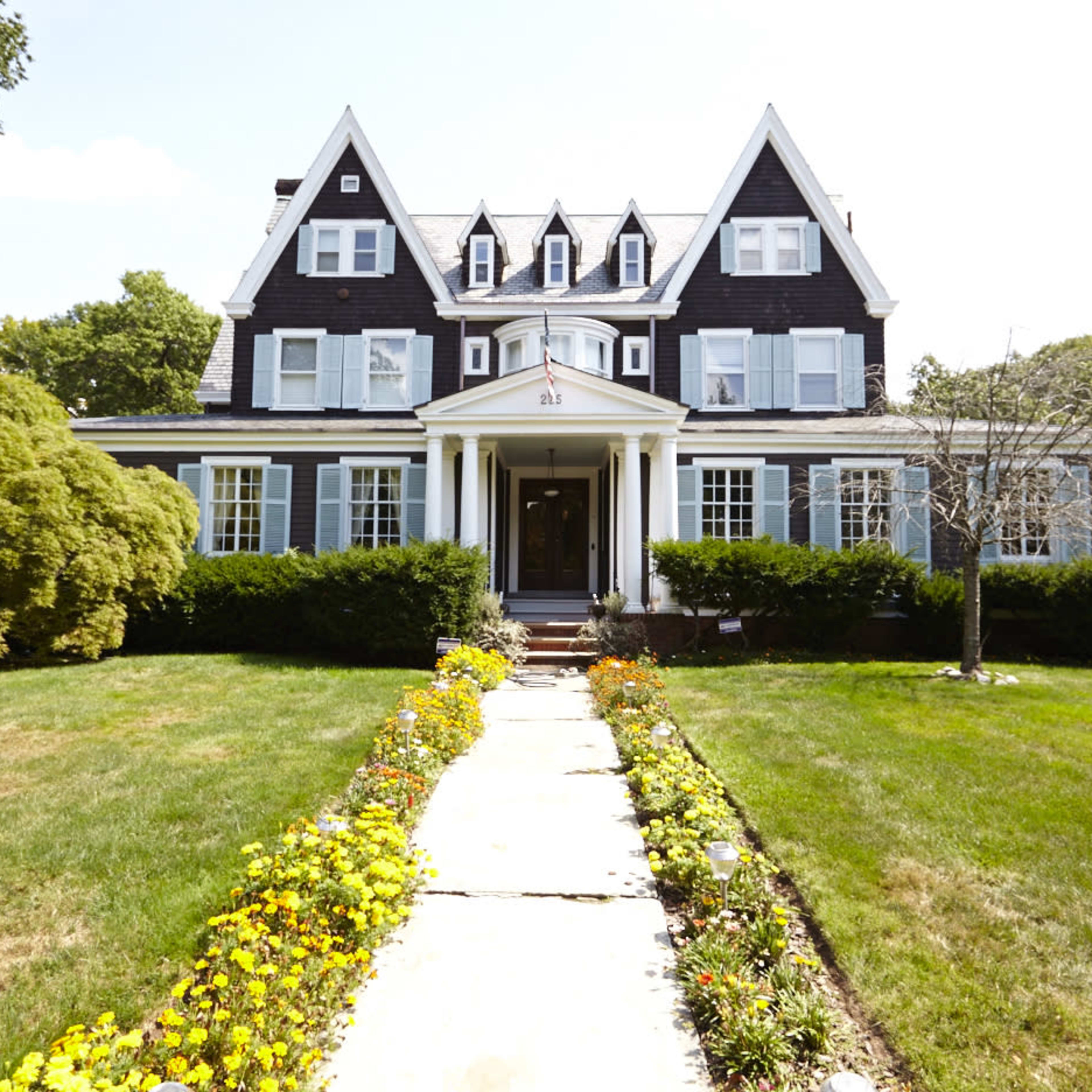 A large, dark-colored house with a steep roof and multiple gables is set behind a well-maintained front yard and pathway lined with colorful flowers.