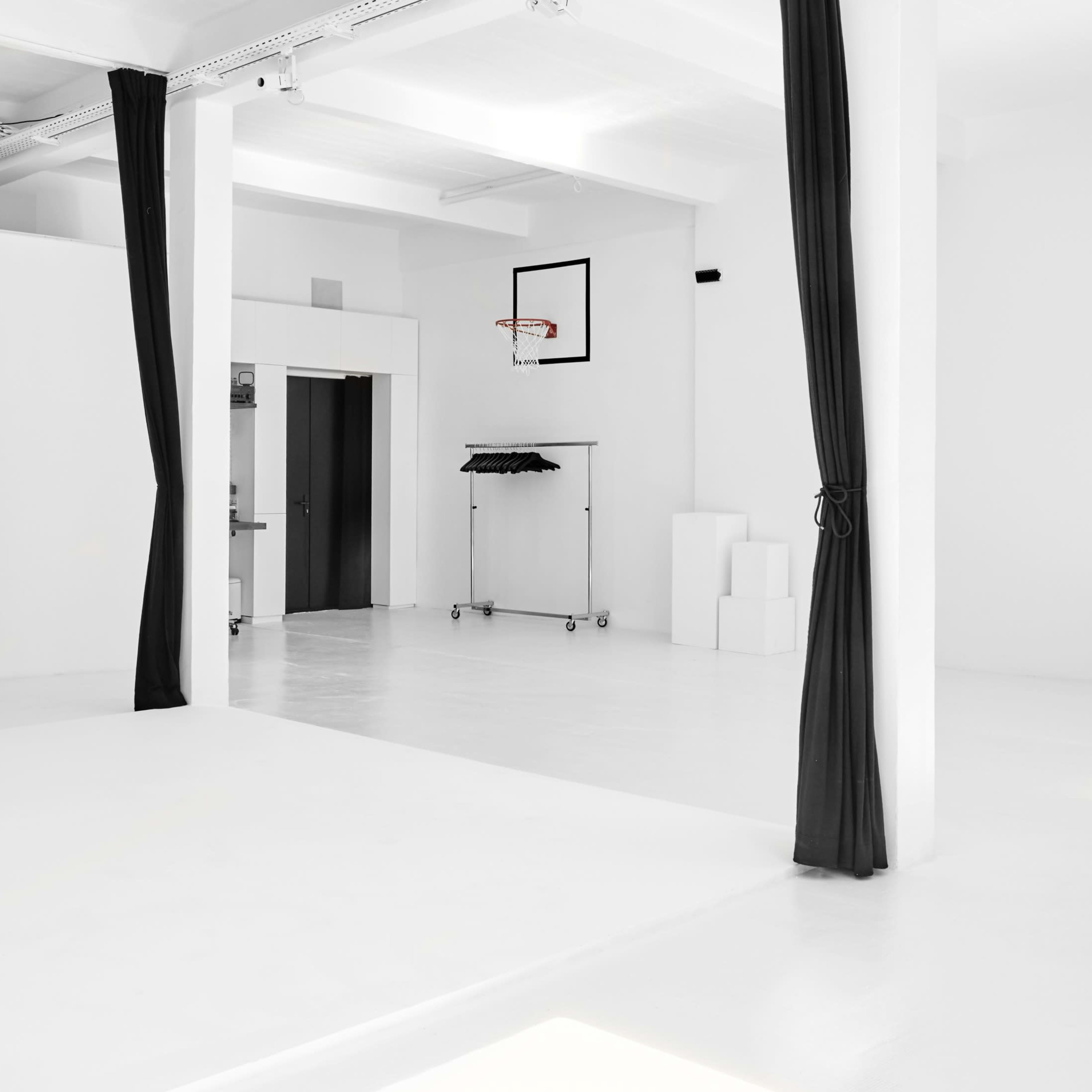 A spacious, minimalist interior with white walls and a kitchenette, featuring a basketball hoop and a clothing rack in one corner.