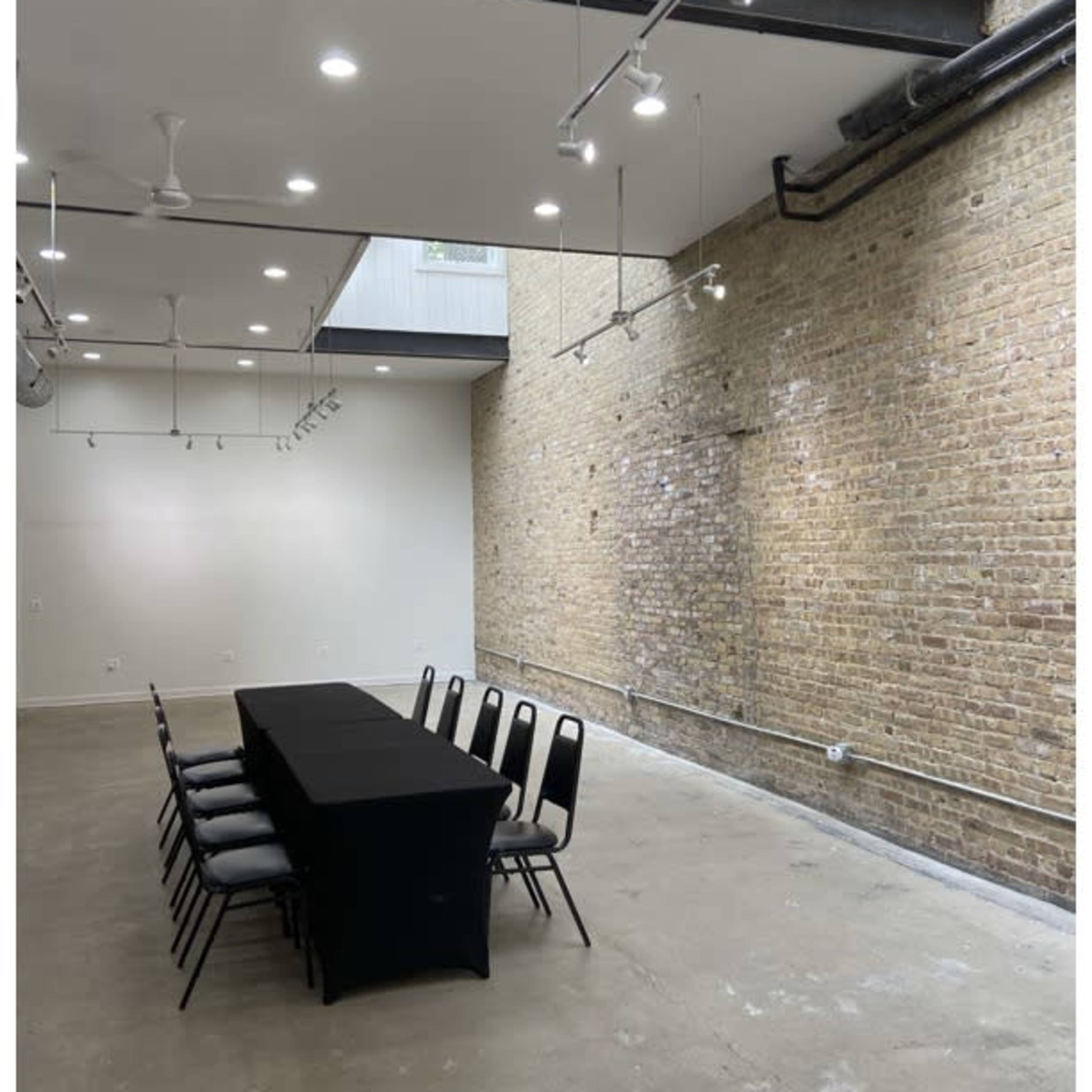 A long black table with chairs is set up in an industrial-style room featuring exposed brick walls and ceiling lights.